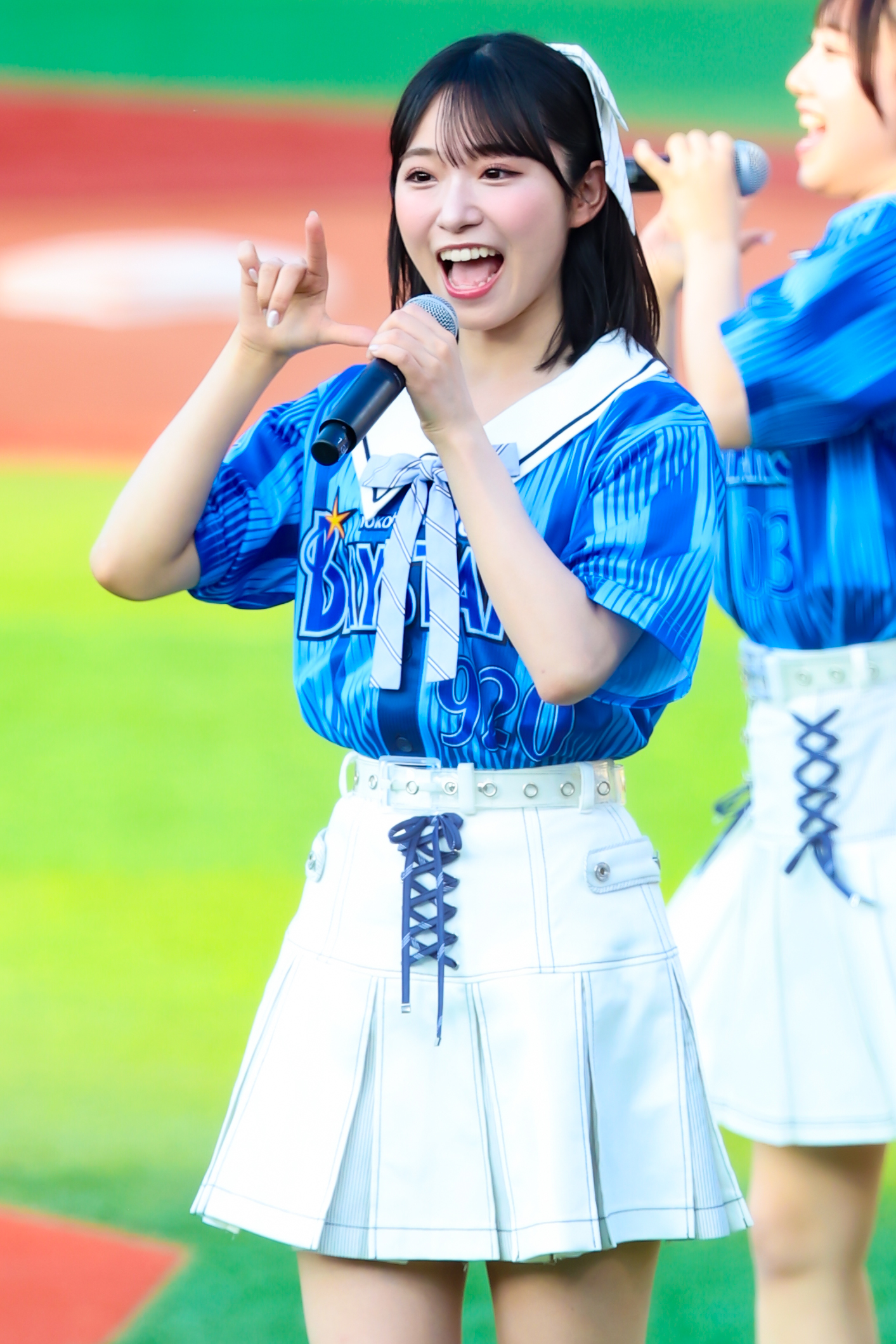A performer from AKB48 wearing a blue shirt and white skirt enthusiastically sings into a microphone while making a hand gesture, with another performer visible in the background.