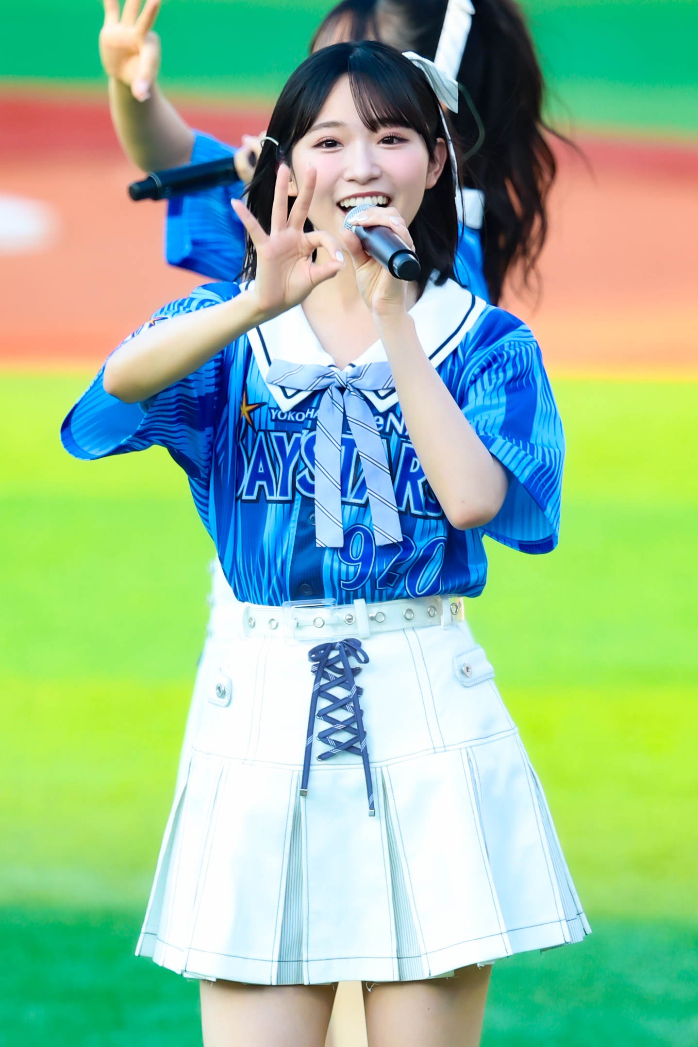 A member of AKB48 wearing a blue jersey and a white skirt, smiling and holding a microphone while performing. The background features a green field.