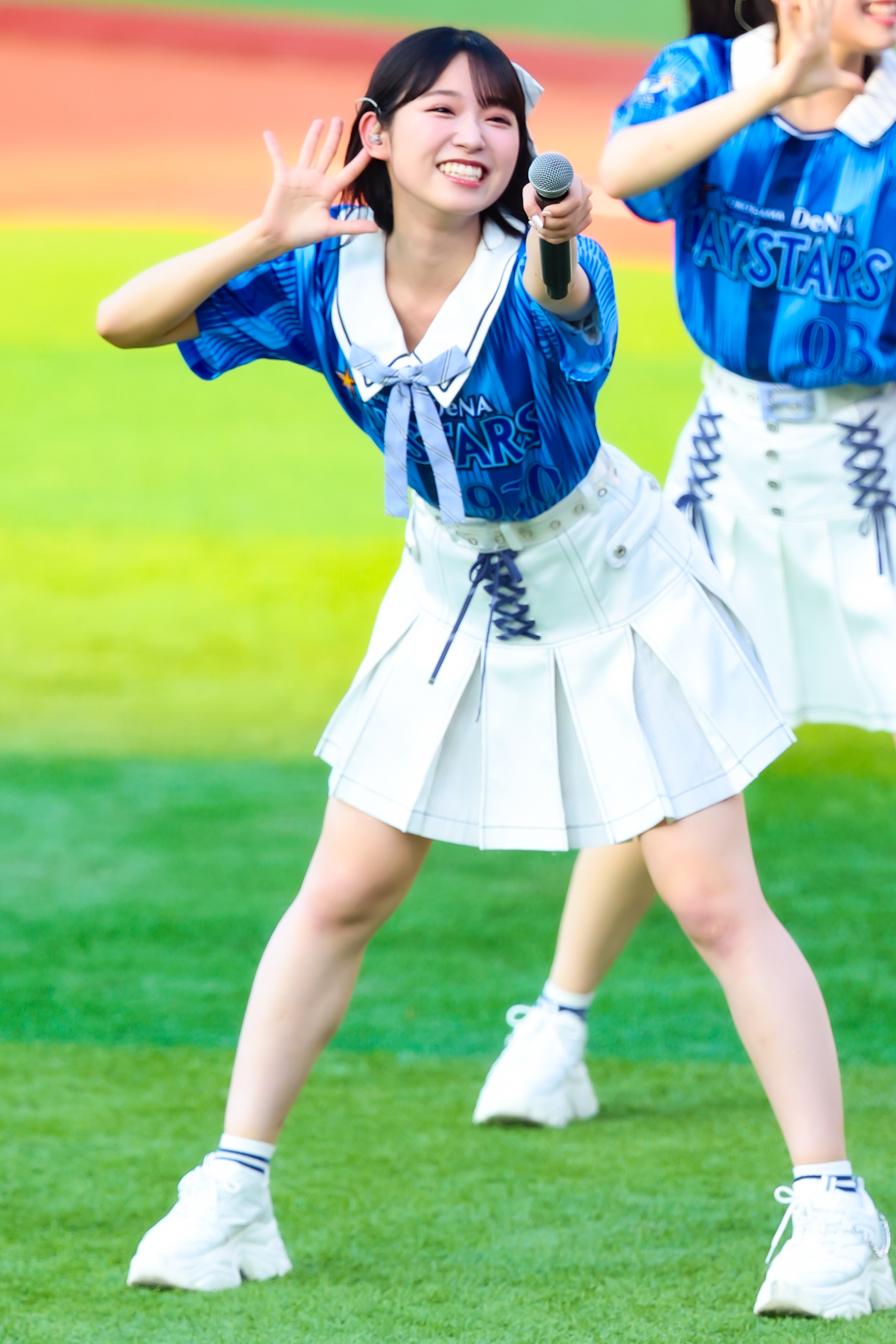 A young performer wearing a blue shirt with 'DeNA BAYSTARS' printed on it, smiling and holding a microphone while dancing on a green field.