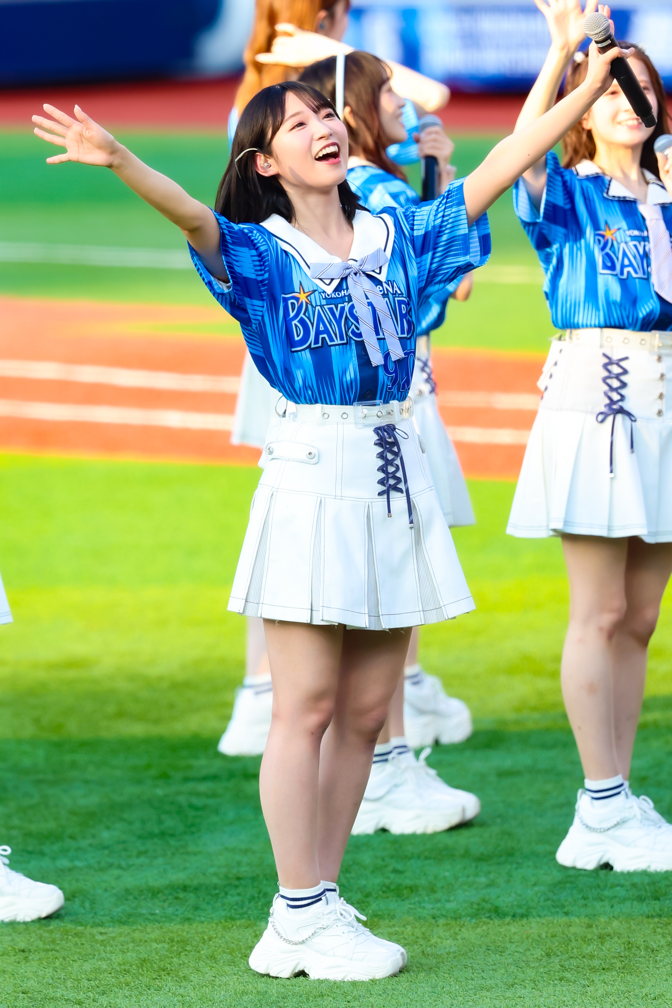 A performer from AKB48 wearing a blue team jersey and a white skirt, joyfully singing with arms outstretched on a grassy field.