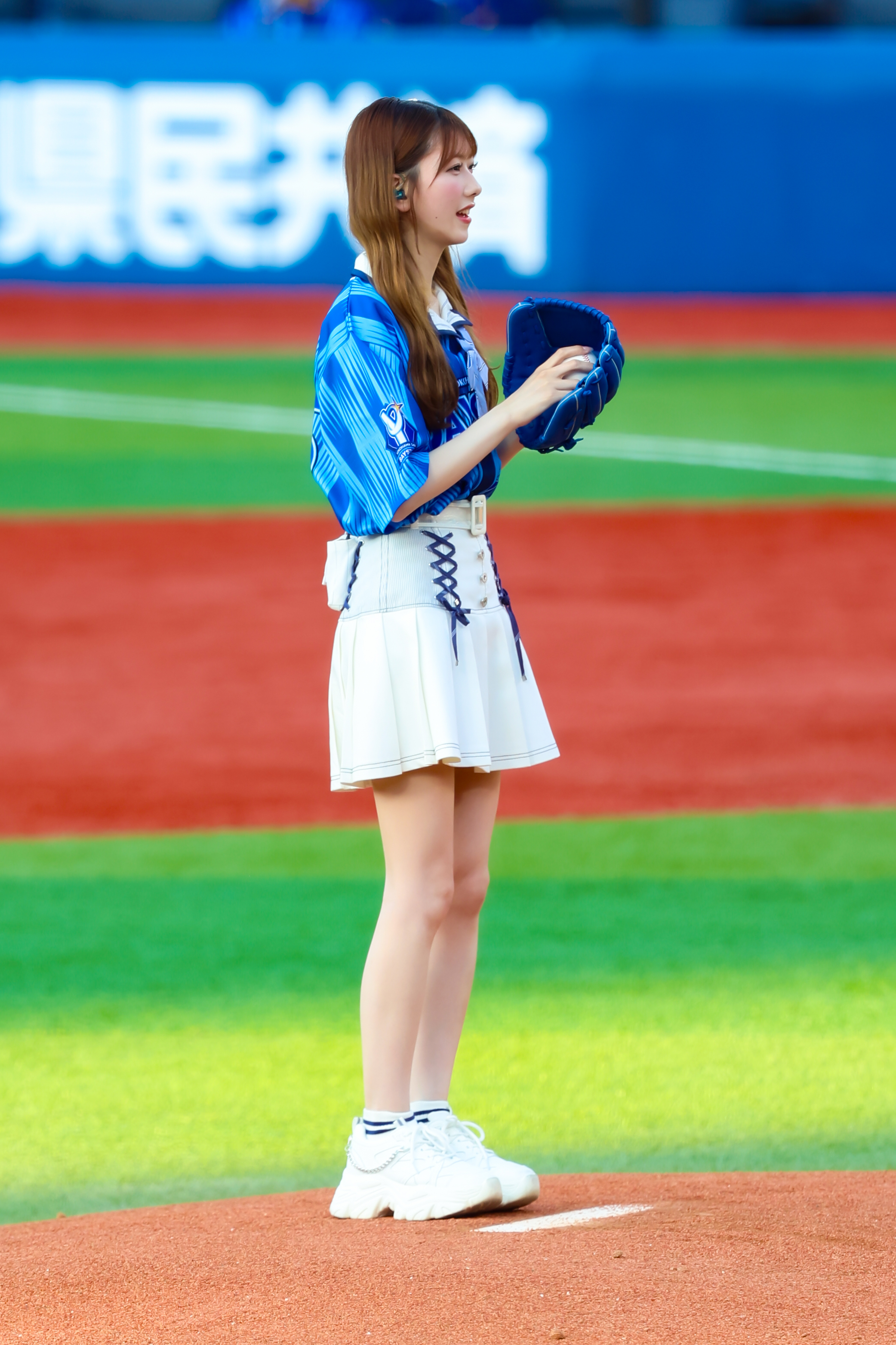 A female performer wearing a blue sports jersey and white skirt stands on a baseball field holding a blue glove, preparing for a pitch.