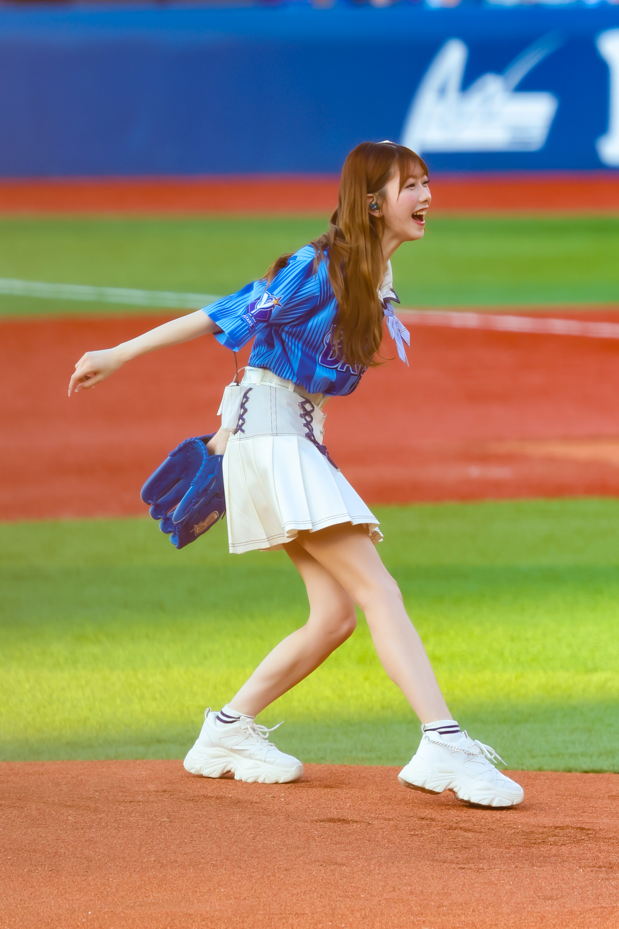 A performer from AKB48 is smiling and running on a baseball field, wearing a blue top and white skirt, while holding a glove.