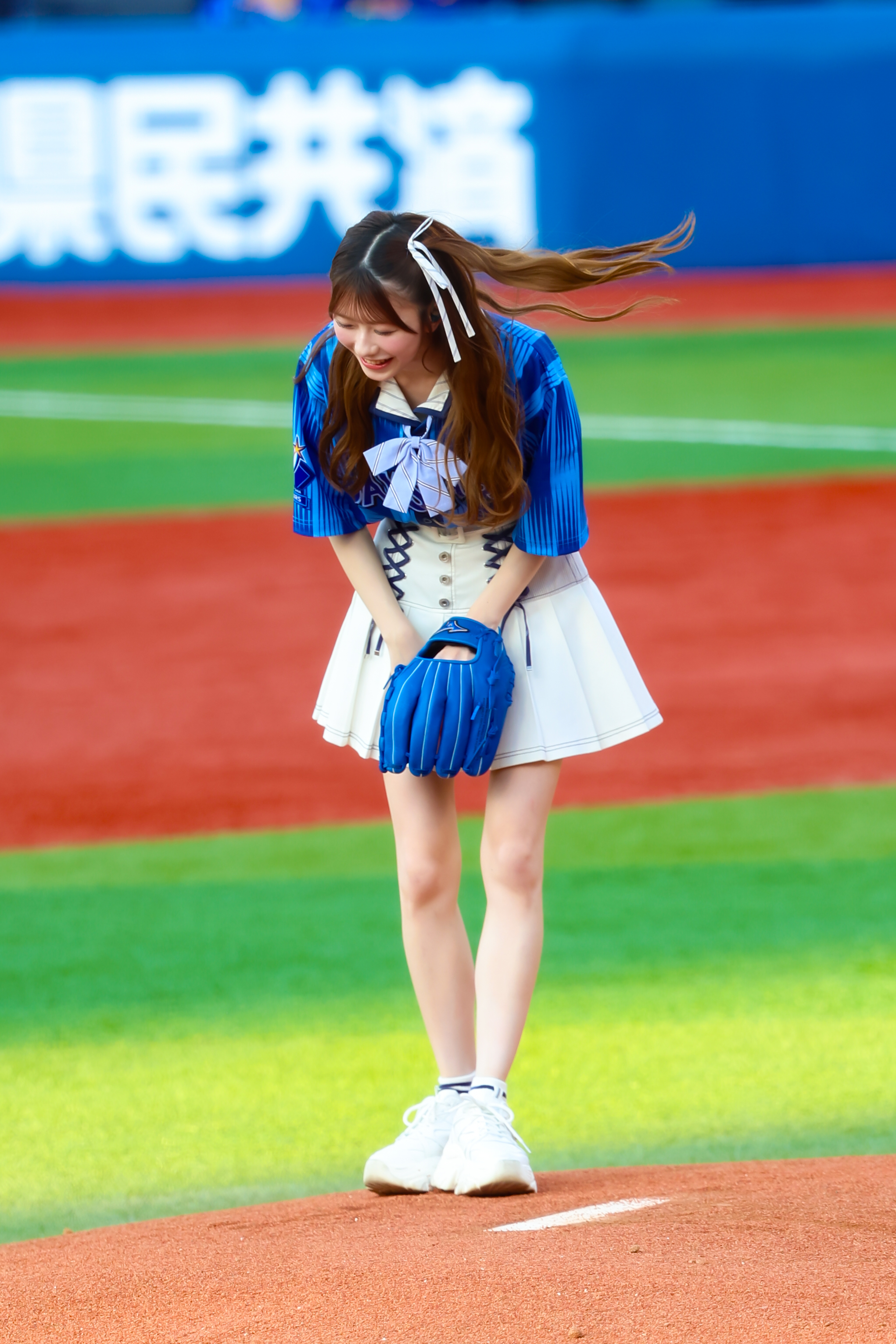A girl wearing a blue jersey and white skirt is preparing to pitch on a baseball field, wearing a blue glove and smiling.