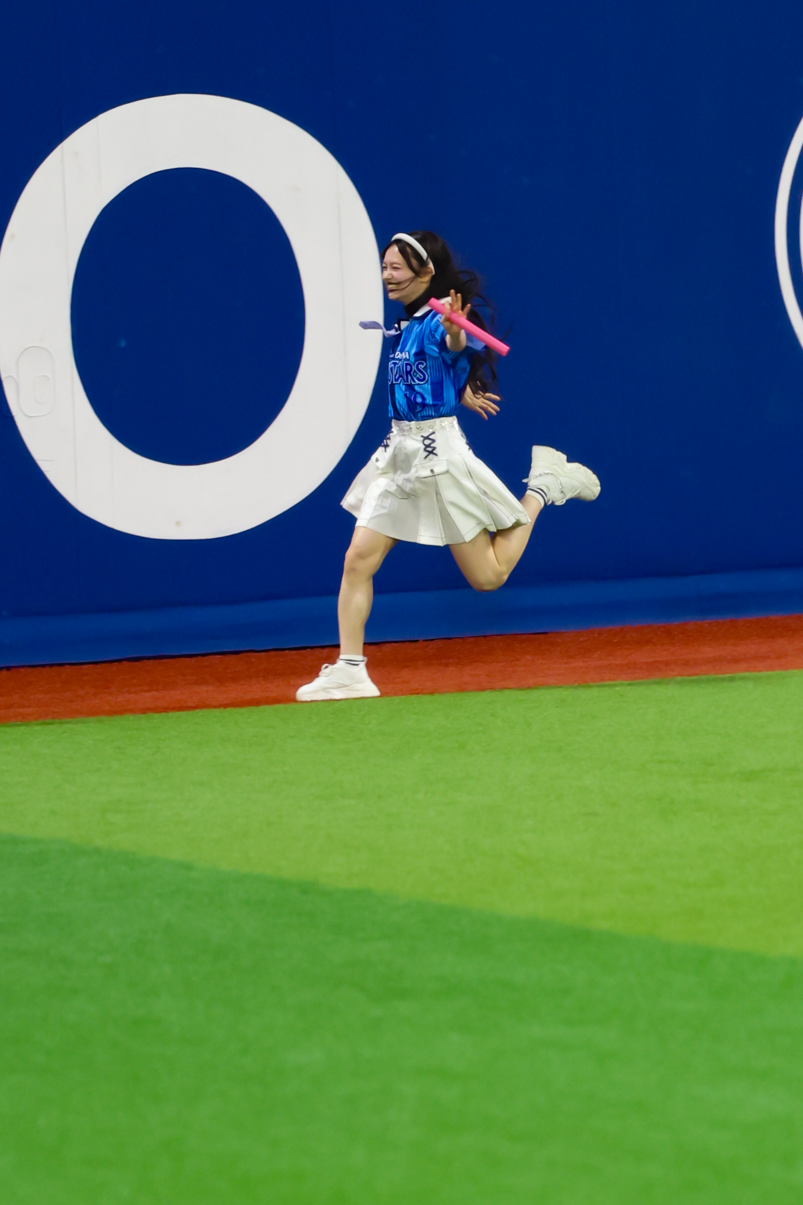 A performer in a blue and white outfit runs energetically on a green turf field, holding a pink baton, with a large letter 'O' visible in the background.