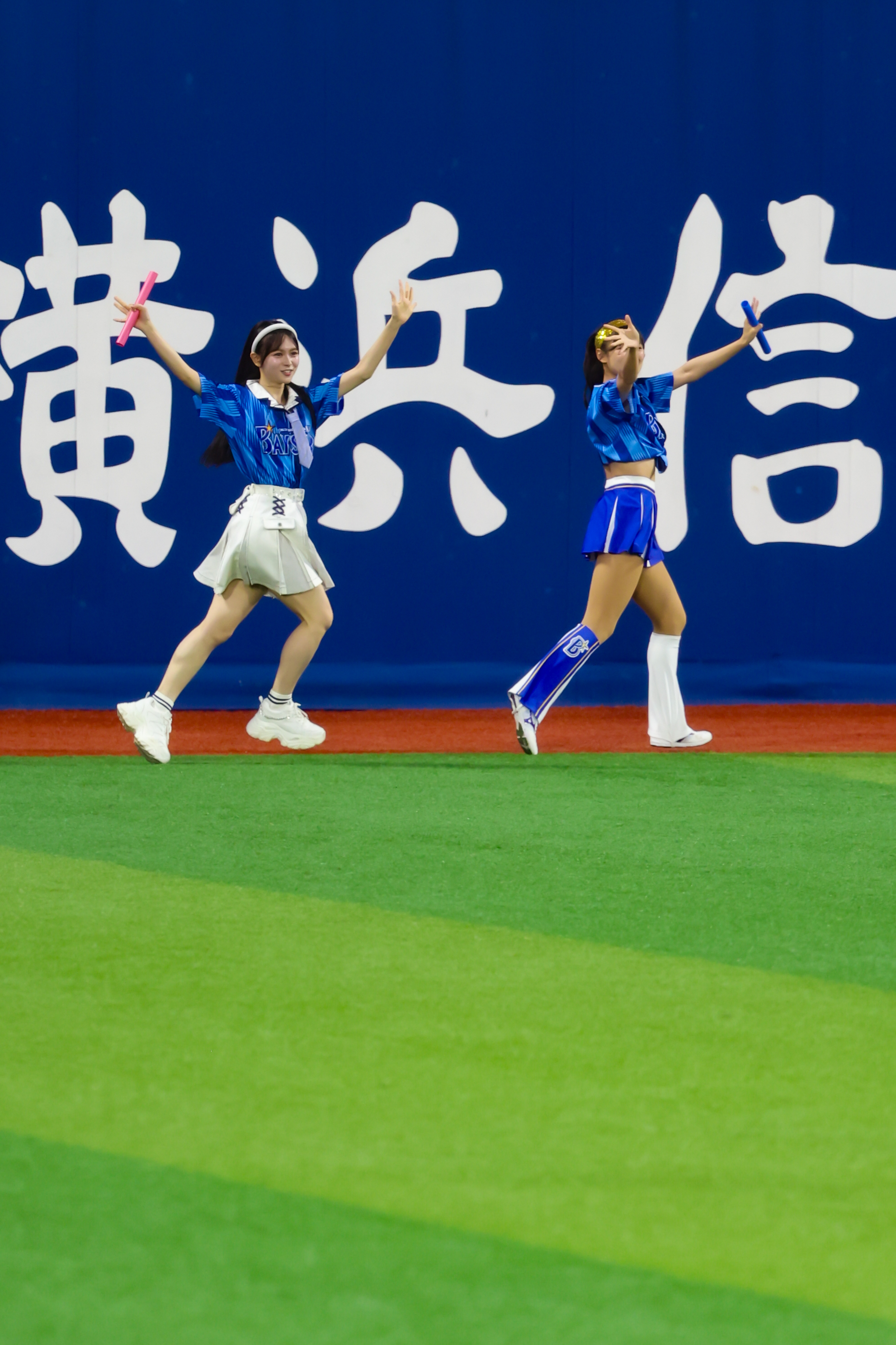 Two performers dancing on a green field with a blue background featuring large white characters. One dancer is wearing a white skirt and the other in a blue outfit.
