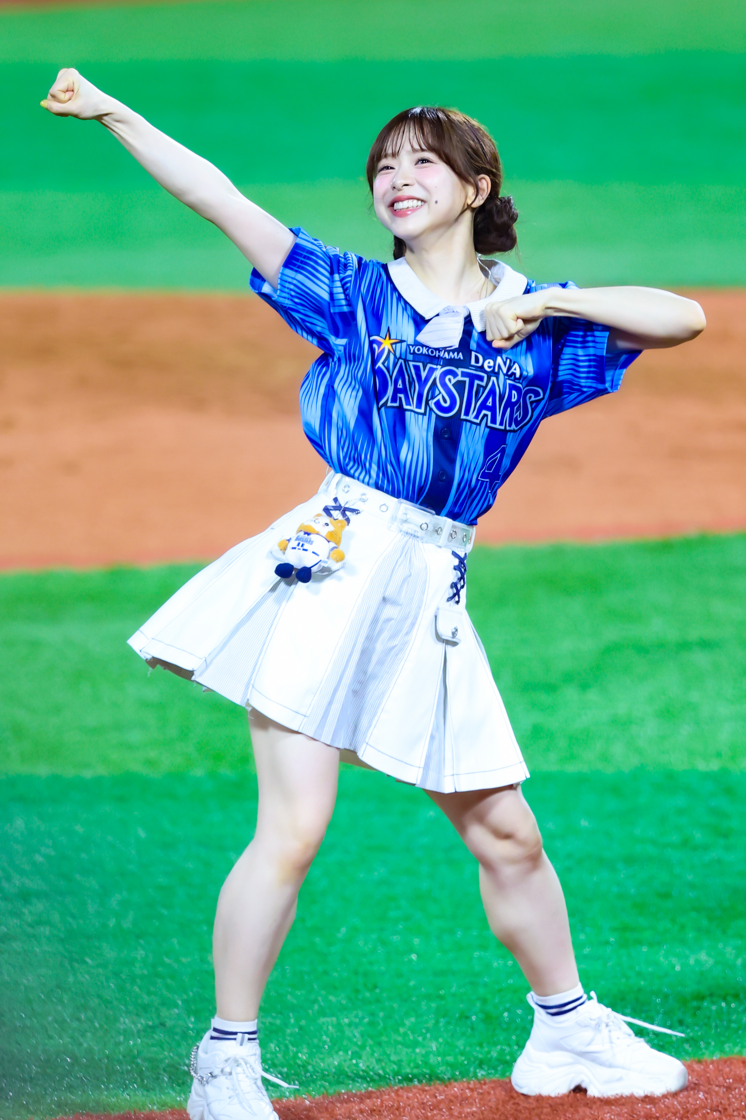 A performer in a blue baseball jersey and white skirt energetically poses with a clenched fist and a big smile on a baseball field.