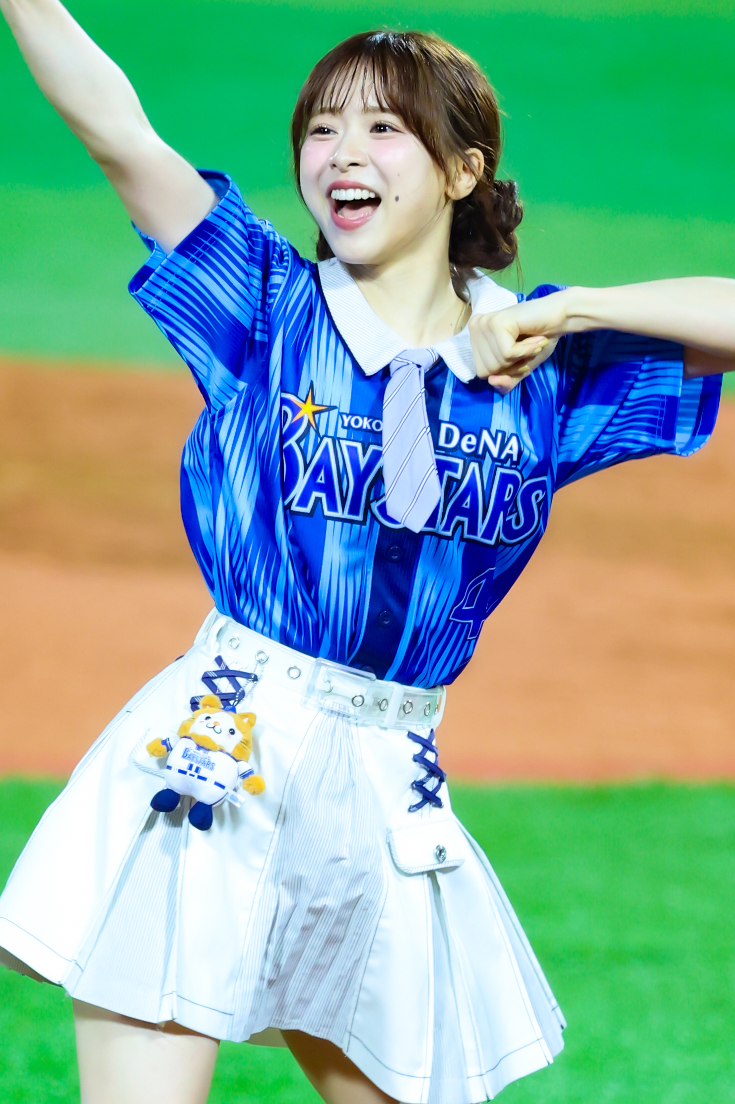 A young woman in a blue Yokohama DeNA BayStars jersey and white skirt, smiling and cheering with her right arm raised. A small plush toy is attached to her waist.