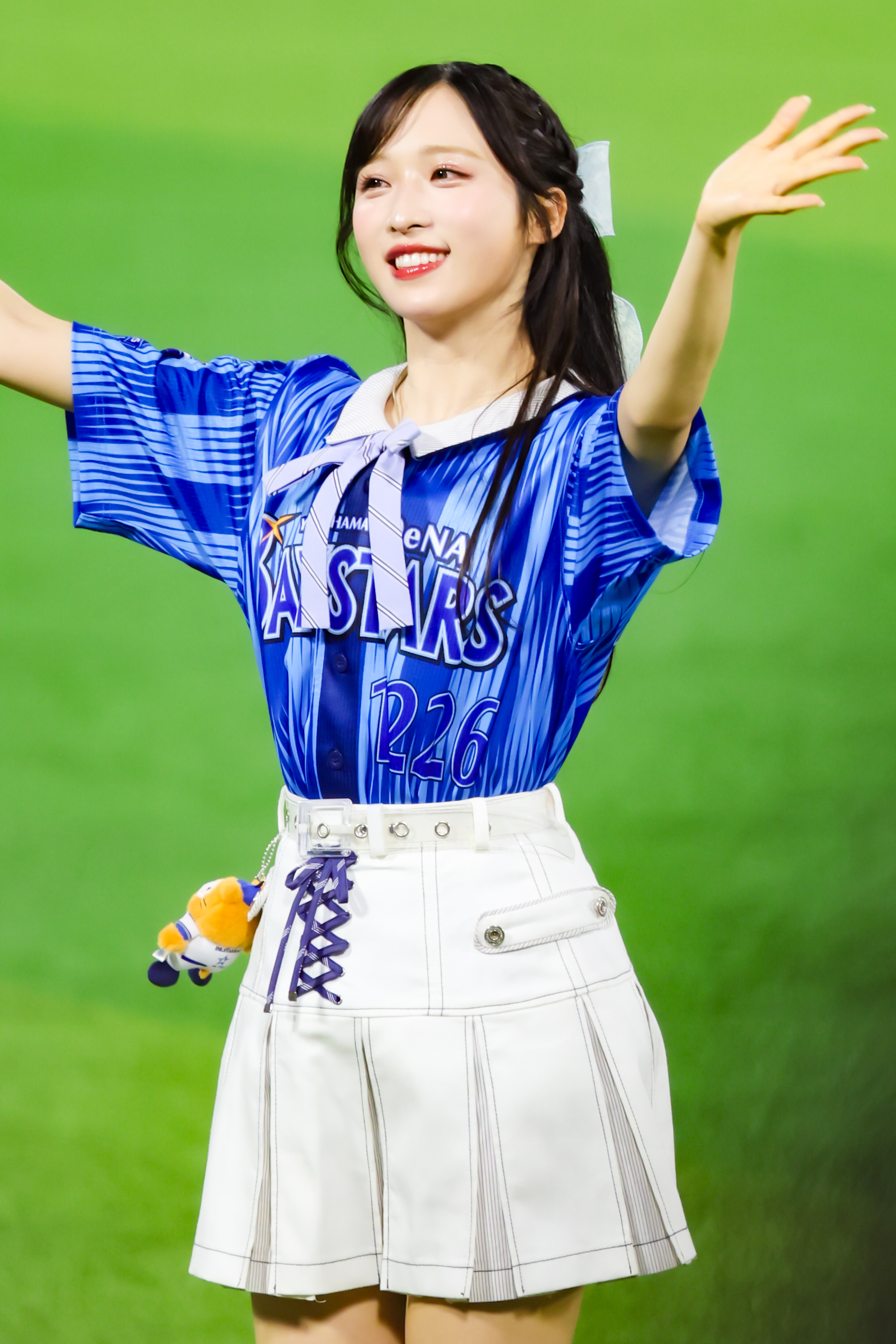 A performer wearing a blue sports jersey with 'Yokohama BayStars' branding and a white skirt is cheering with arms raised in a lively manner on a baseball field.