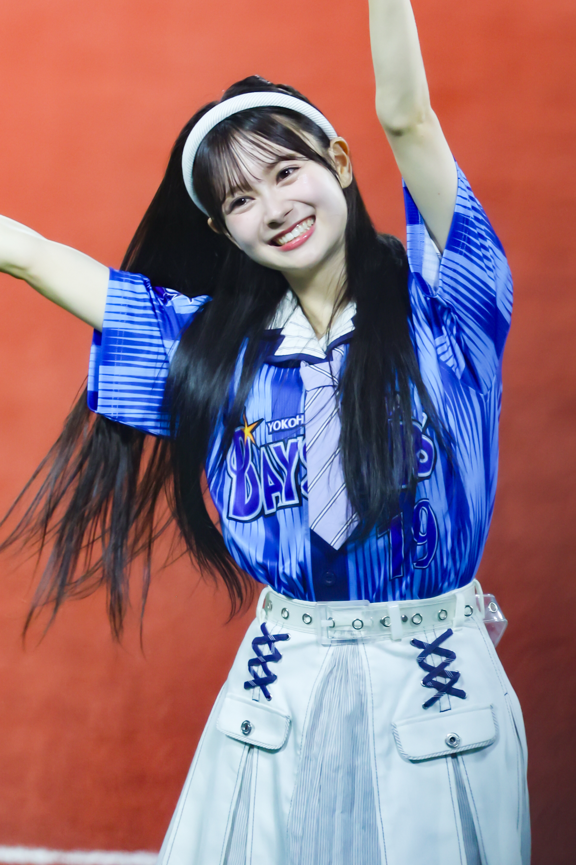 A smiling girl wearing a blue baseball-style shirt with white details and a white skirt, enthusiastically raising her arms against a vibrant orange background.