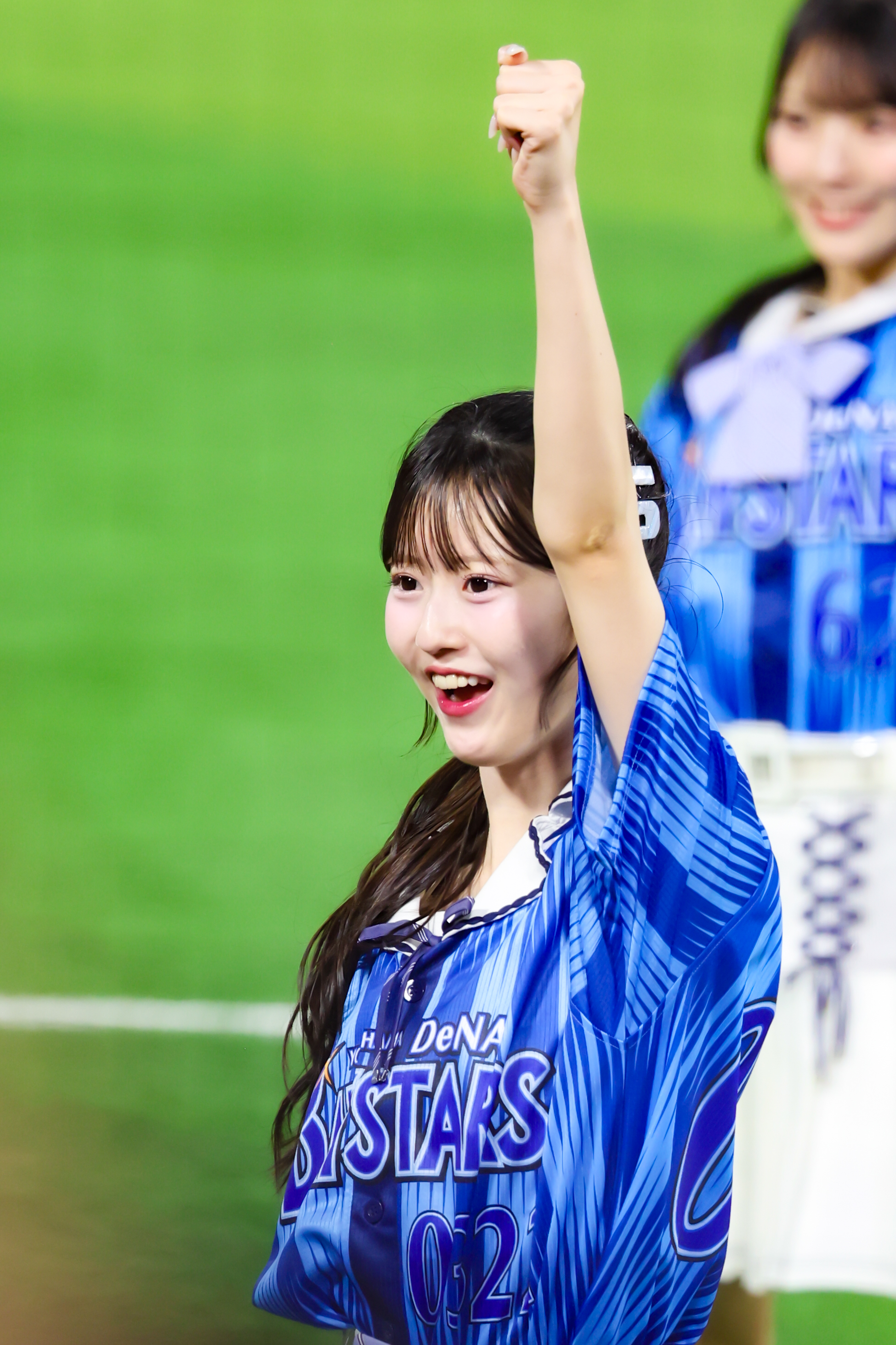 A young woman dressed in a blue sports jersey raises her fist in celebration while smiling during an outdoor event.