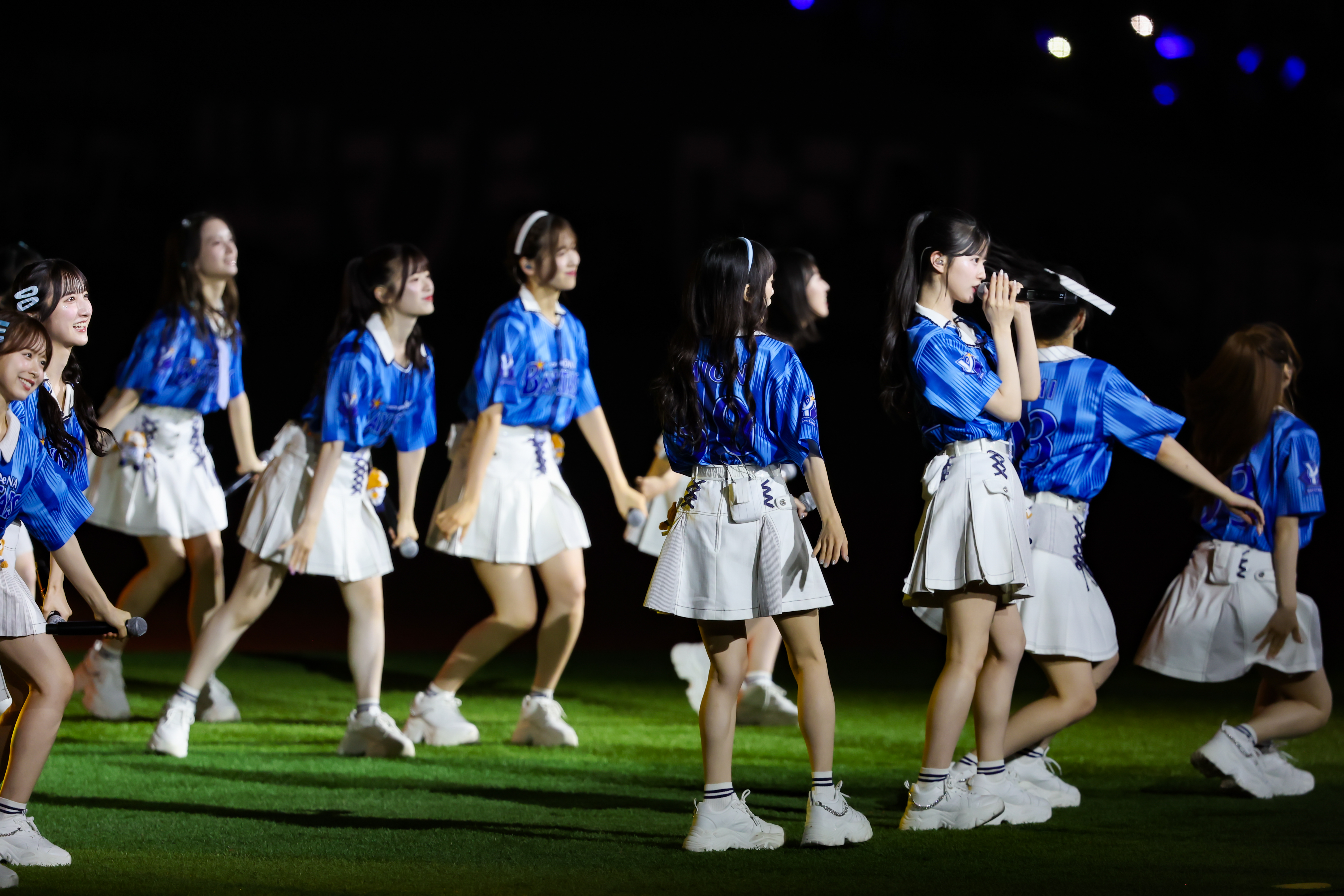 A group of singers from AKB48 performing on stage, wearing blue jerseys and white skirts, engaging with the audience during a concert.