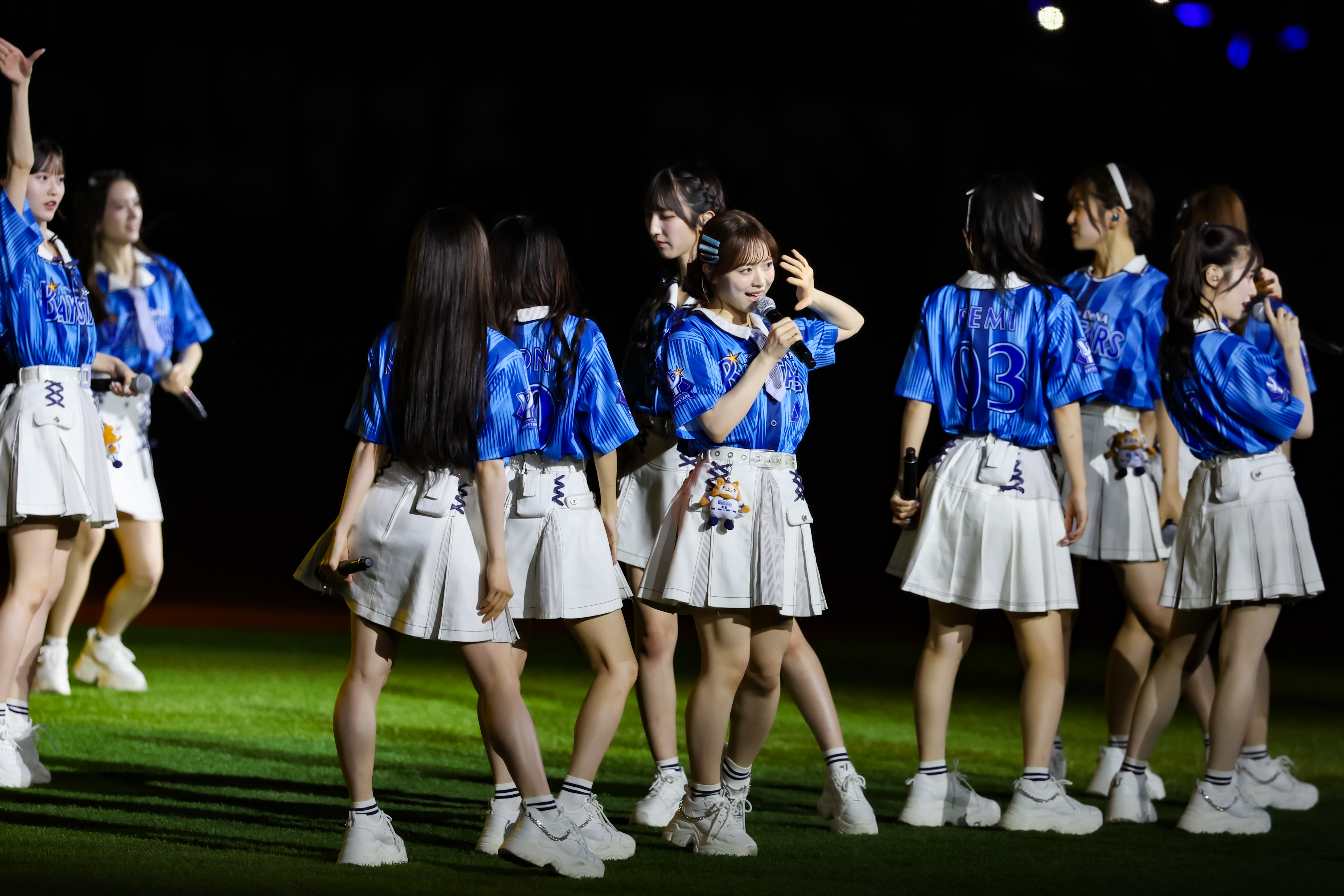 A group of performers from AKB48 in blue and white uniforms on stage. One member is speaking into a microphone, while others are engaged with each other. The scene is illuminated with stage lights, set in a vibrant atmosphere.