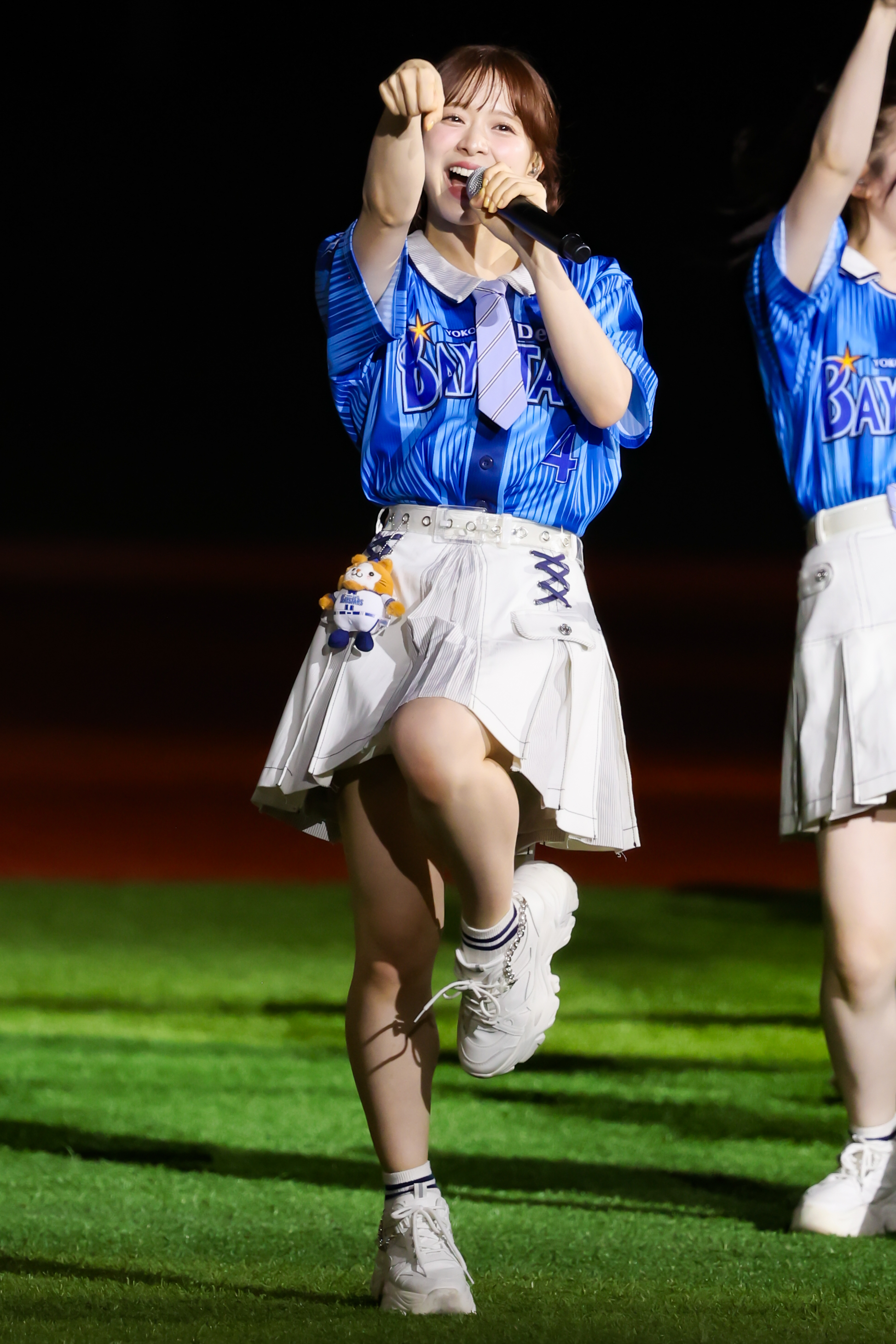 A performer from AKB48 wearing a blue jersey and a white skirt is singing with enthusiasm, pointing at the audience while holding a microphone.