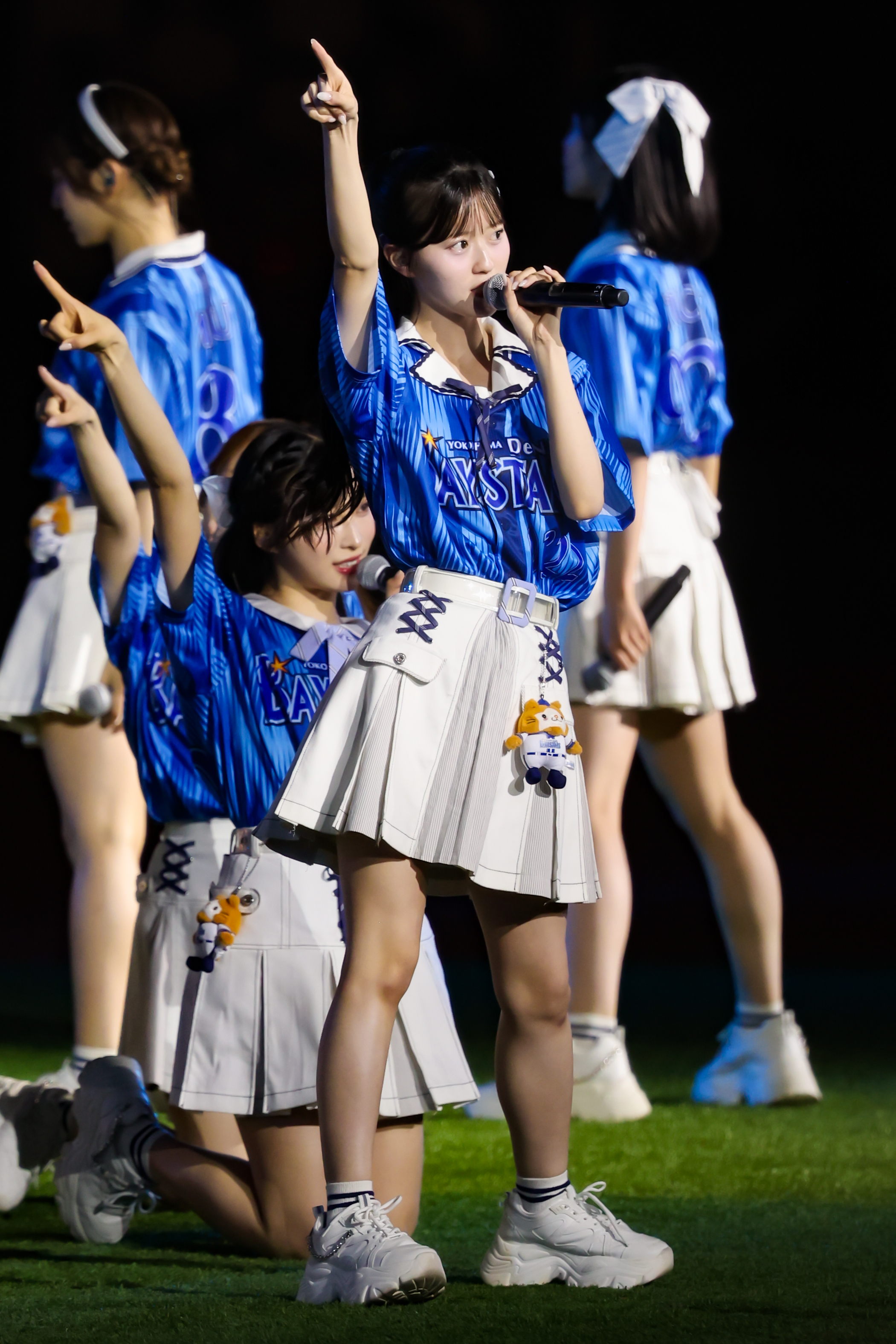 A performer from AKB48 singing on stage, wearing a blue shirt and white skirt, while raising one hand in a pose. Another member is kneeling in front.
