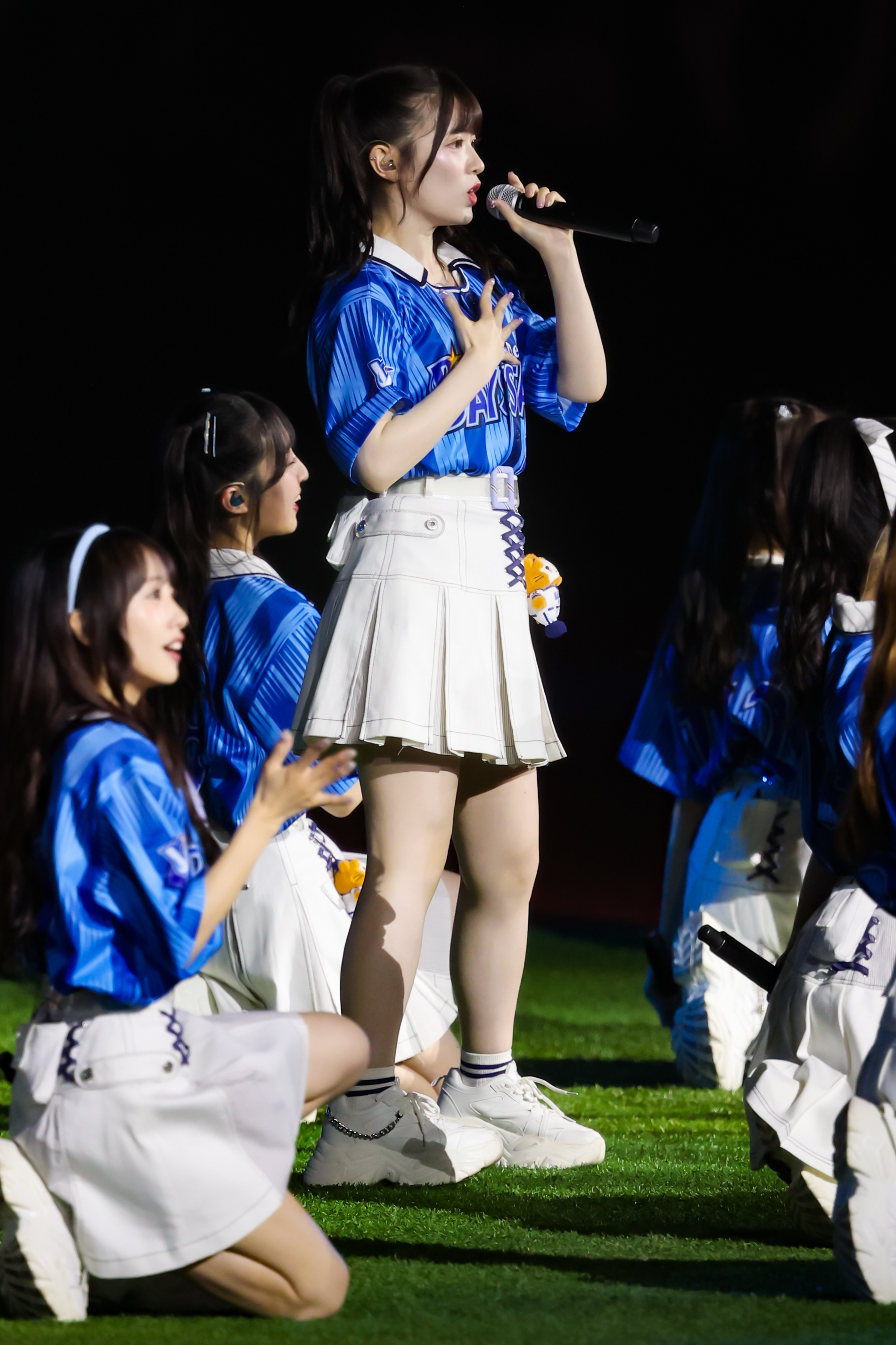 A performer from AKB48 in a blue jersey and white skirt sings into a microphone while surrounded by fellow group members in matching outfits, on stage during a live event.