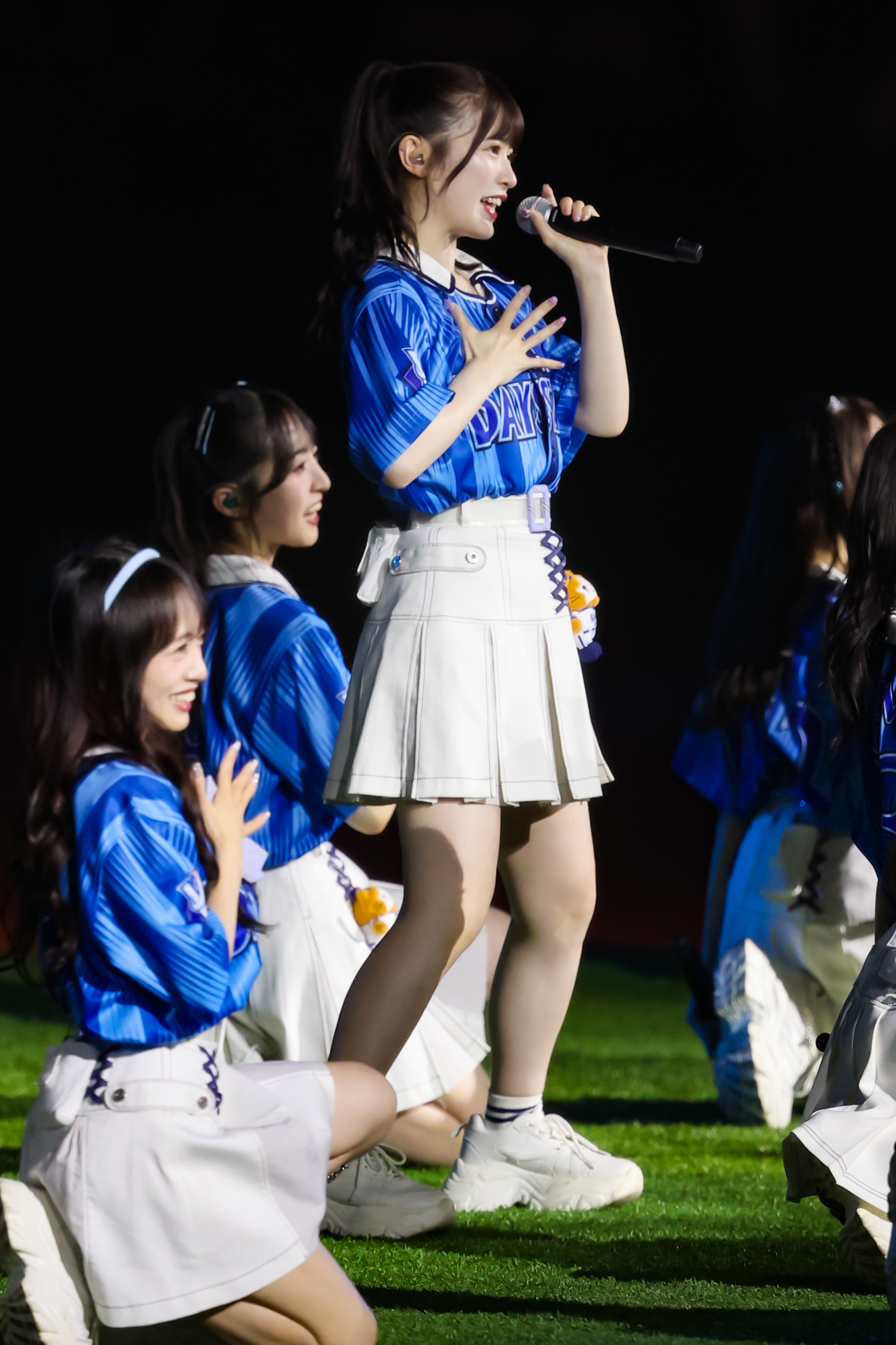 A member of AKB48 performing on stage, wearing a blue jersey and white skirt, holding a microphone with a joyful expression, while other members kneel in the background.