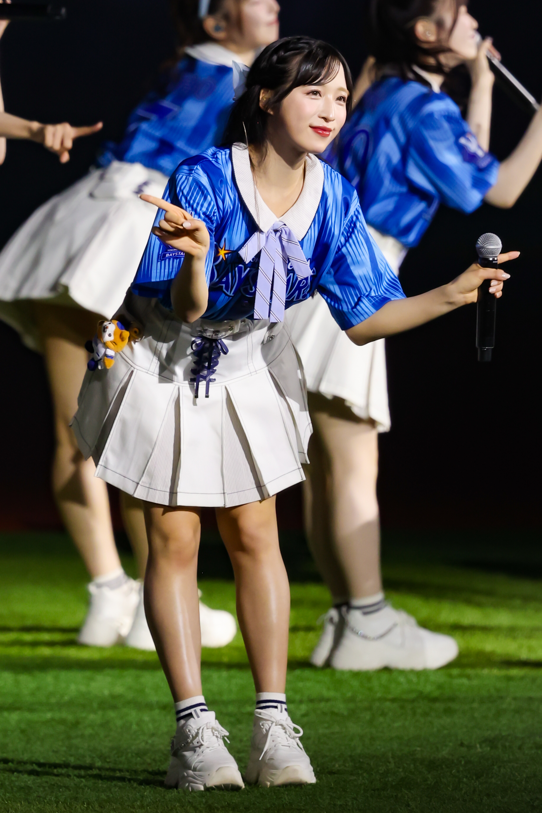 A performer from AKB48 in a blue shirt and white skirt gestures playfully on stage while singing, with other members visible in the background.