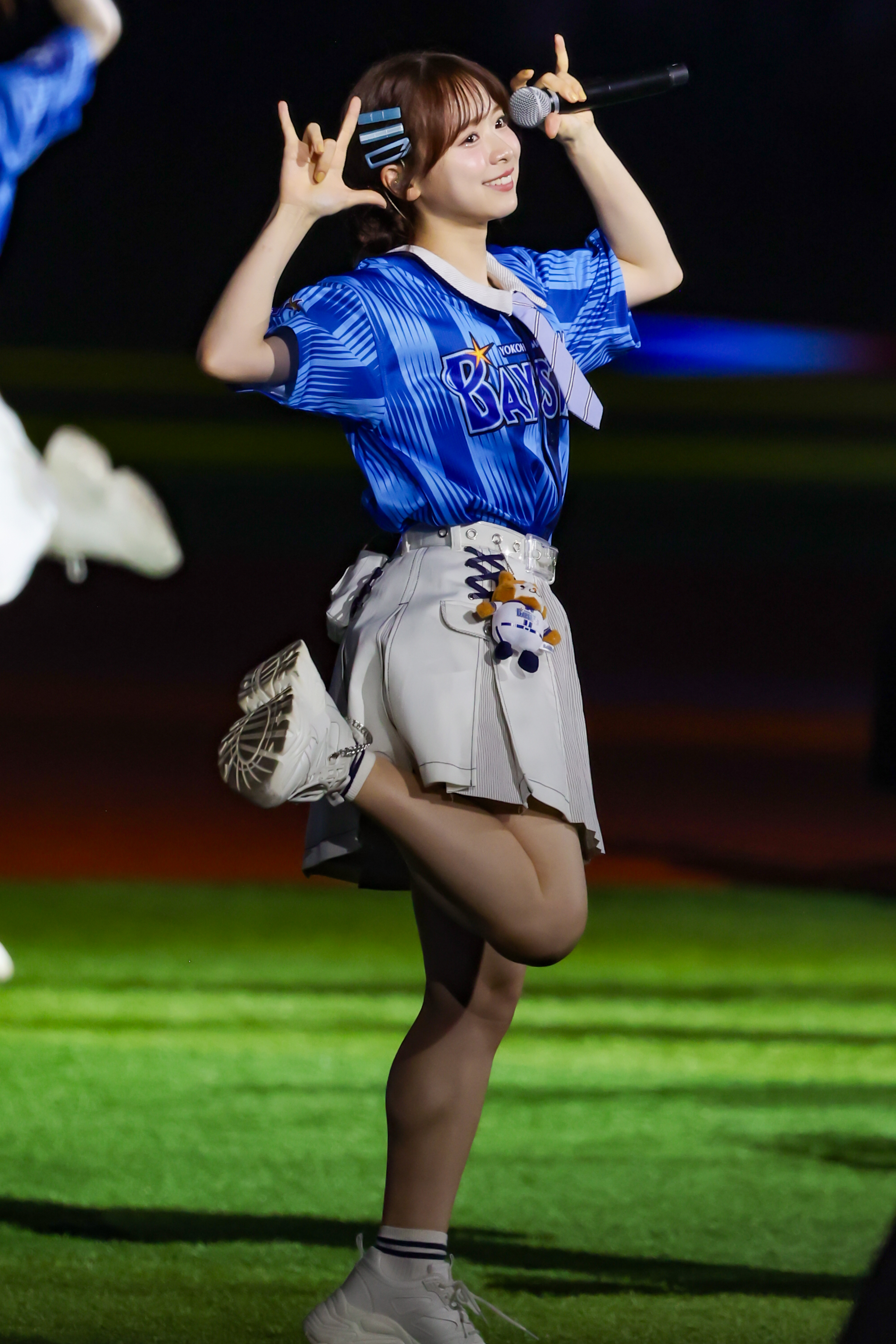 A performer wearing a blue sports jersey and a skirt dances on stage while holding a microphone. She is posing playfully with both hands making gestures, against a lively backdrop.
