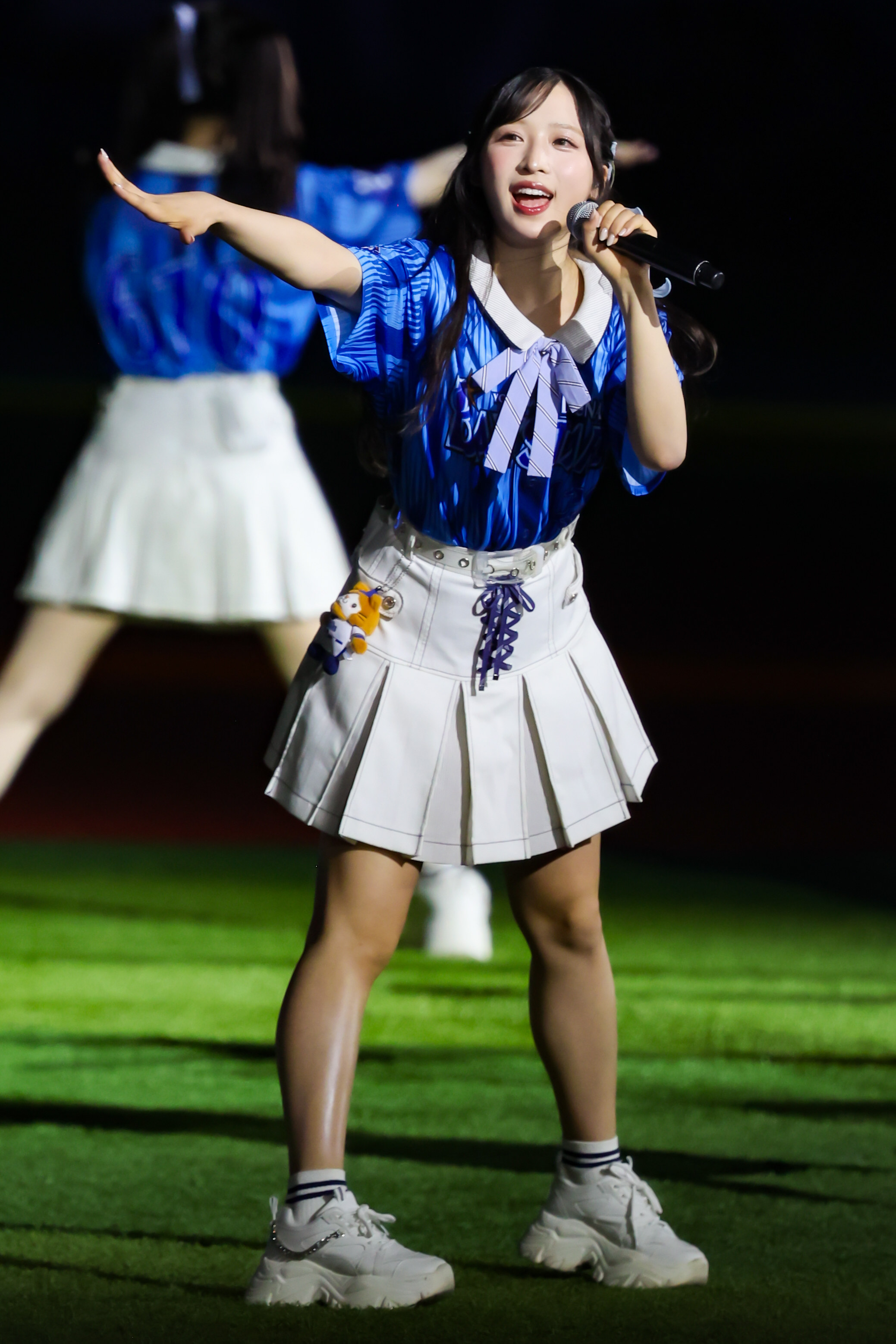A performer energetically singing with a microphone, wearing a blue striped shirt and a white skirt, during a live event on a green field.