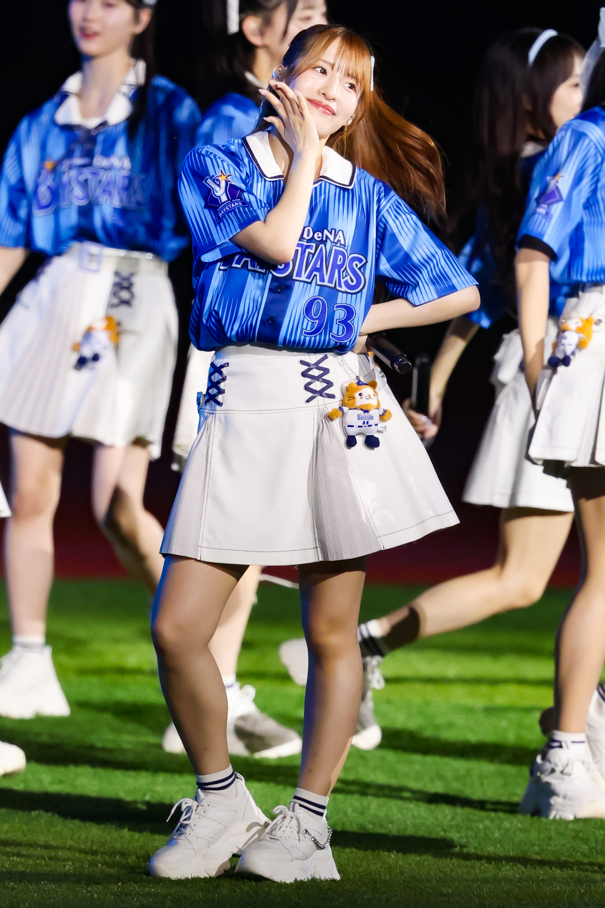 A performer from AKB48 is seen dancing on stage wearing a blue shirt with 'DeNA STARS' printed on it and a white skirt. She is playfully posing with a smile, and there are other dancers in the background.