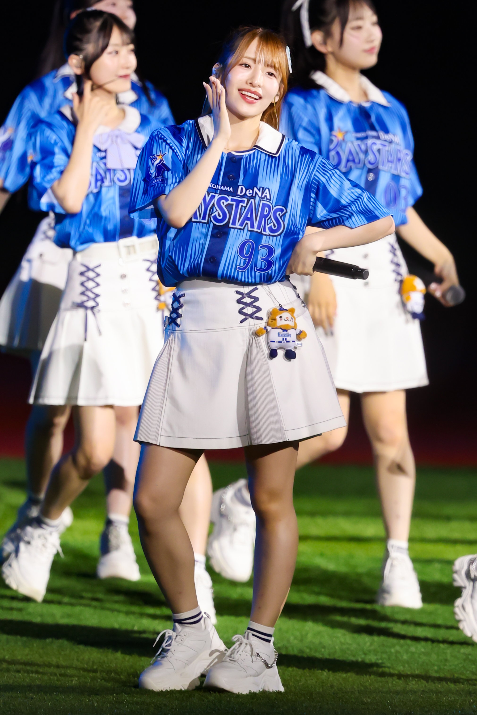 A performer wearing a blue jersey with the number 93 and a white skirt smiles while dancing on stage, surrounded by other performers in matching outfits.