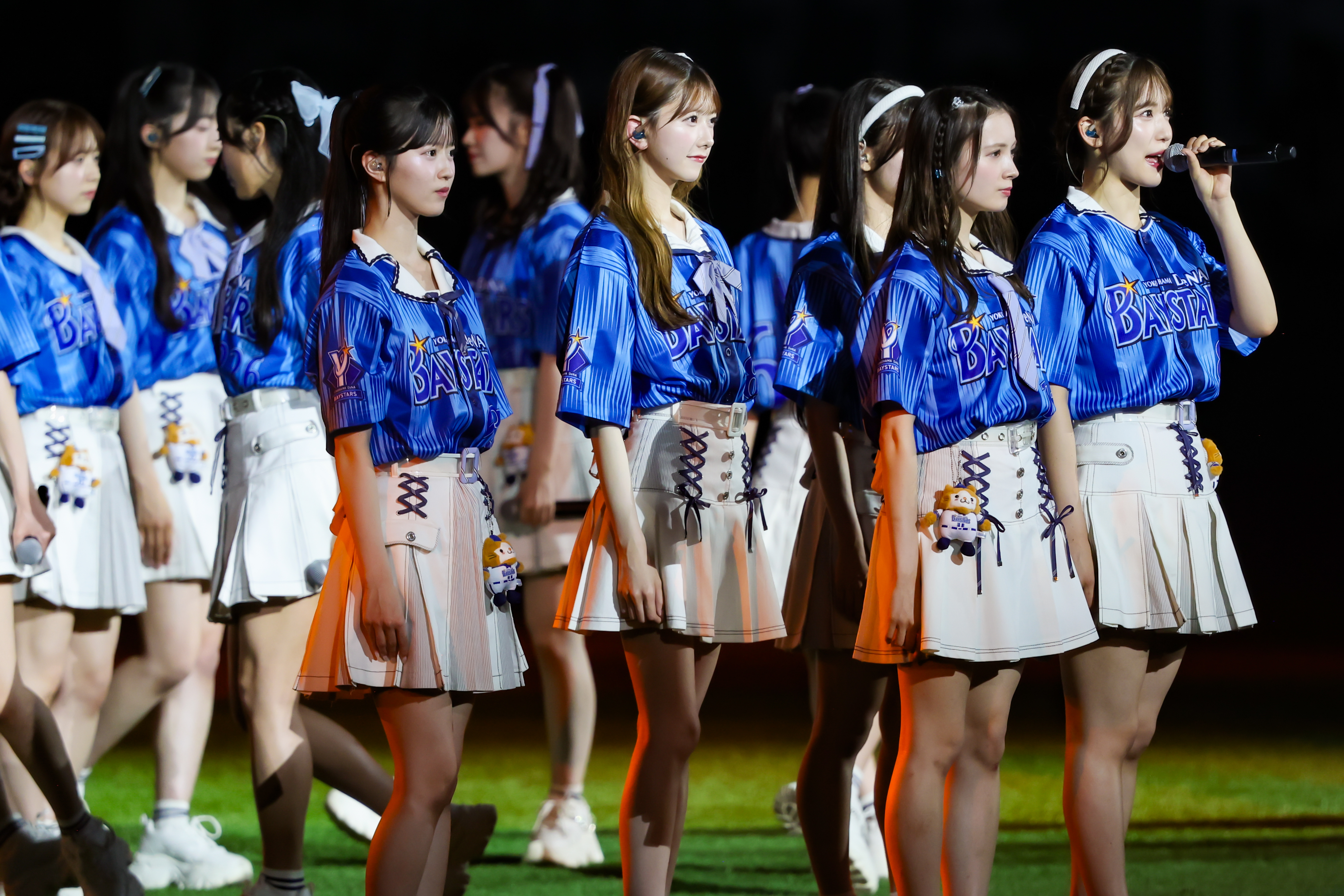 A group of performers from AKB48 wearing blue jerseys and white skirts, standing on stage during a live event. One member is speaking into a microphone while others are focused on their performance.