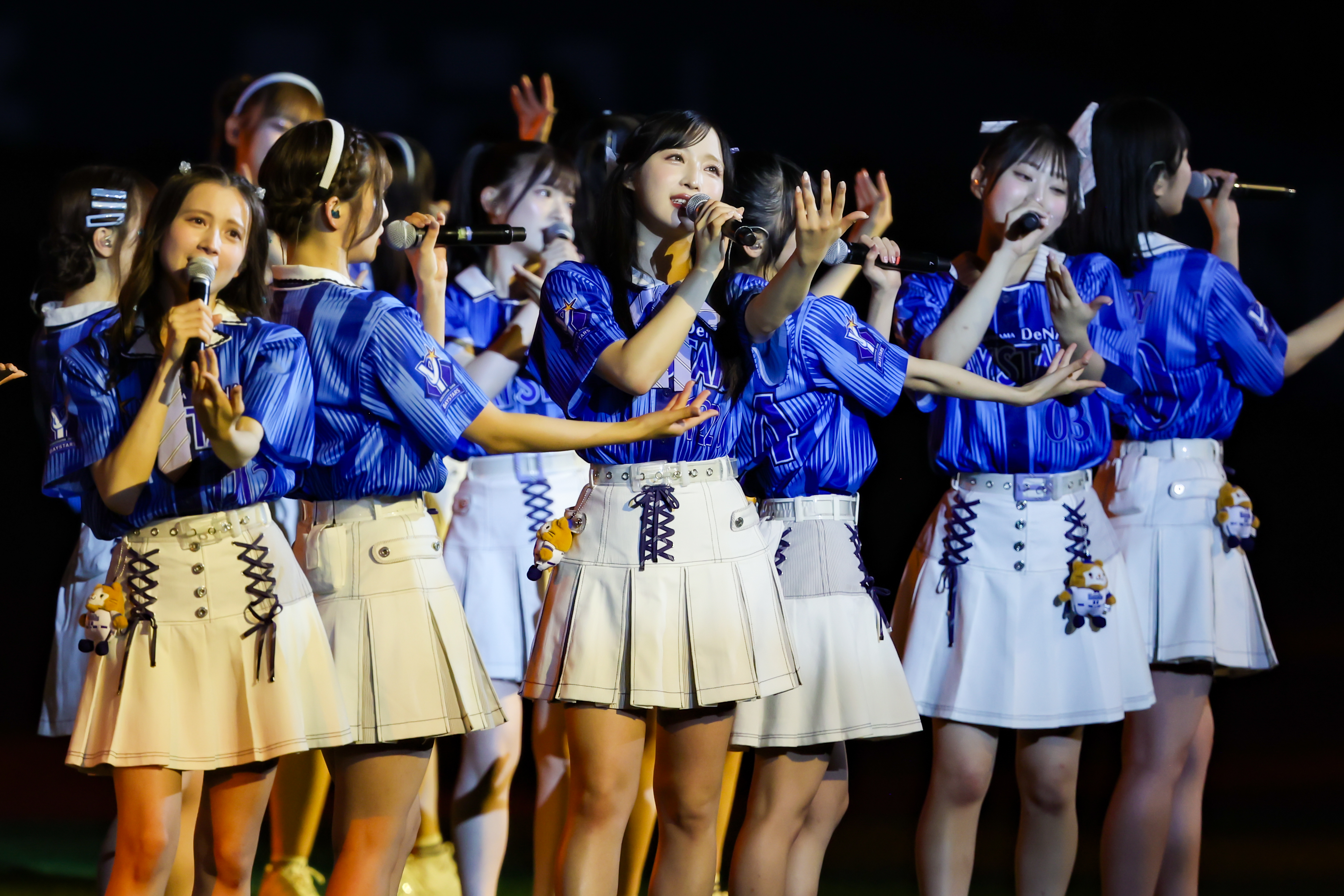 A group of performers from AKB48 in blue and white outfits, singing and engaging with the audience on stage during a concert.