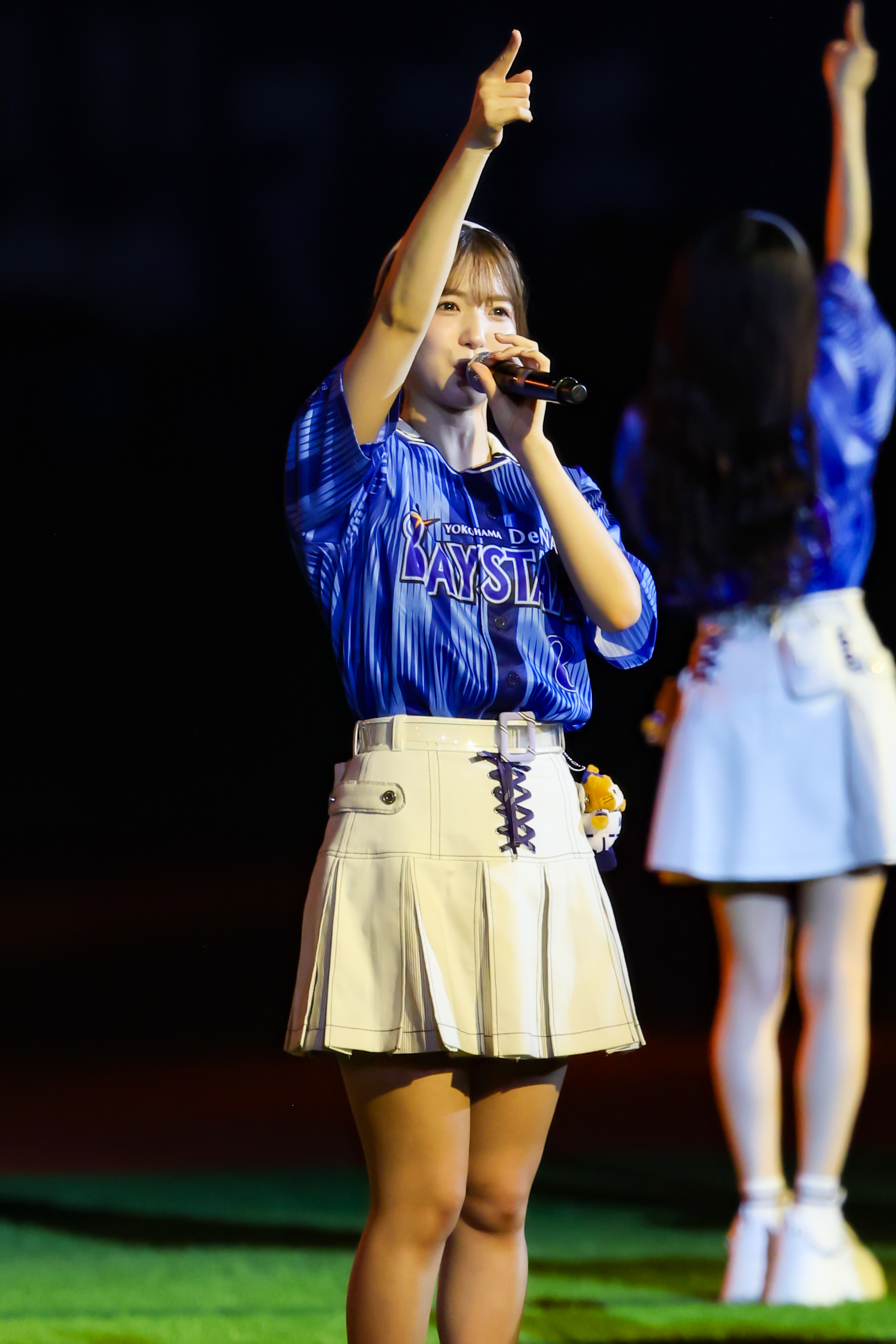 A young female performer wearing a blue shirt and white skirt sings into a microphone while raising her hand in a cheering gesture. Another performer wearing a light-colored skirt is slightly blurred in the background.