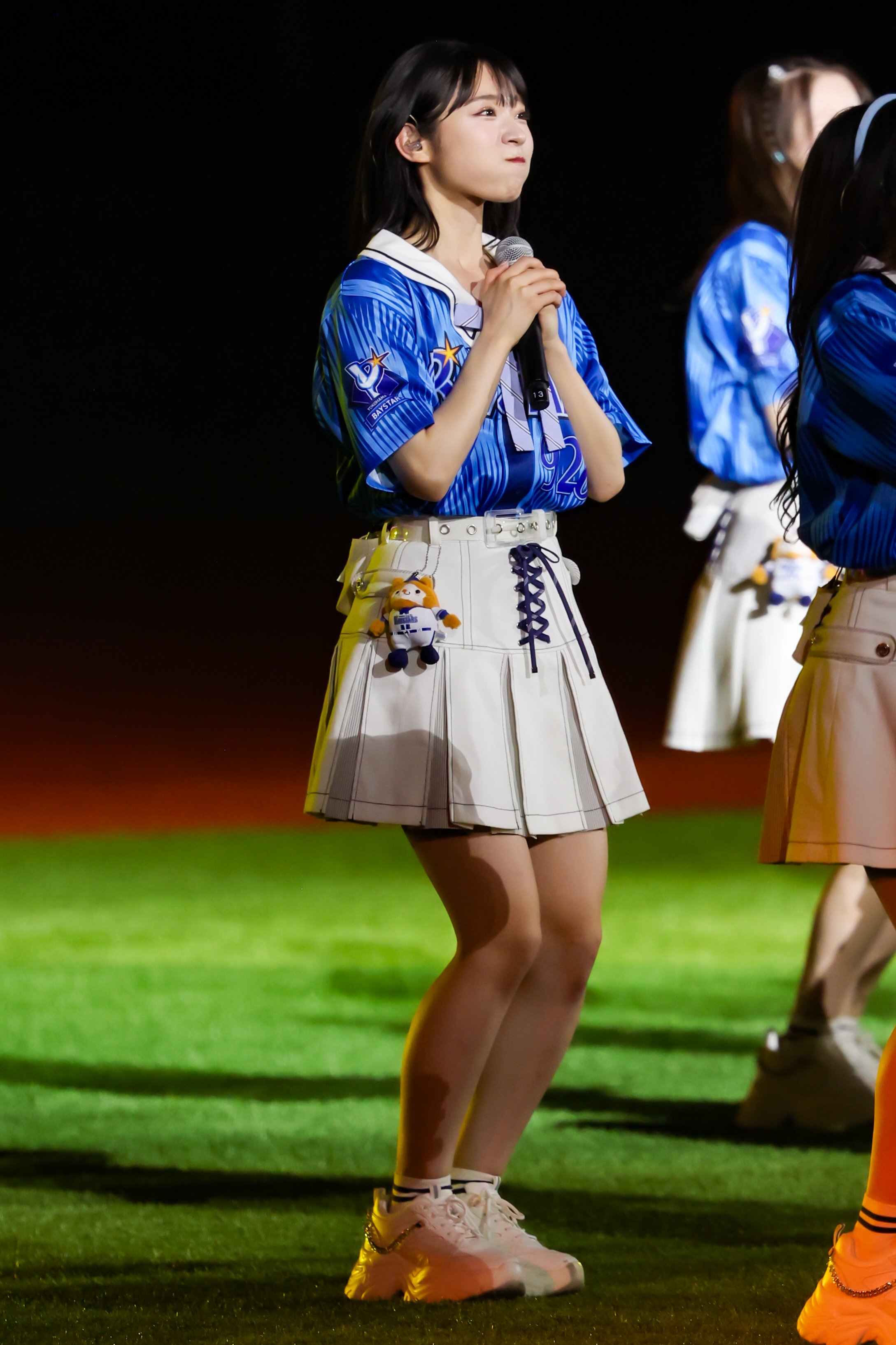A member of AKB48 performing on stage, wearing a blue and white outfit with a belt and holding a microphone. She has a stuffed toy attached to her skirt and is standing on green grass under stage lights.