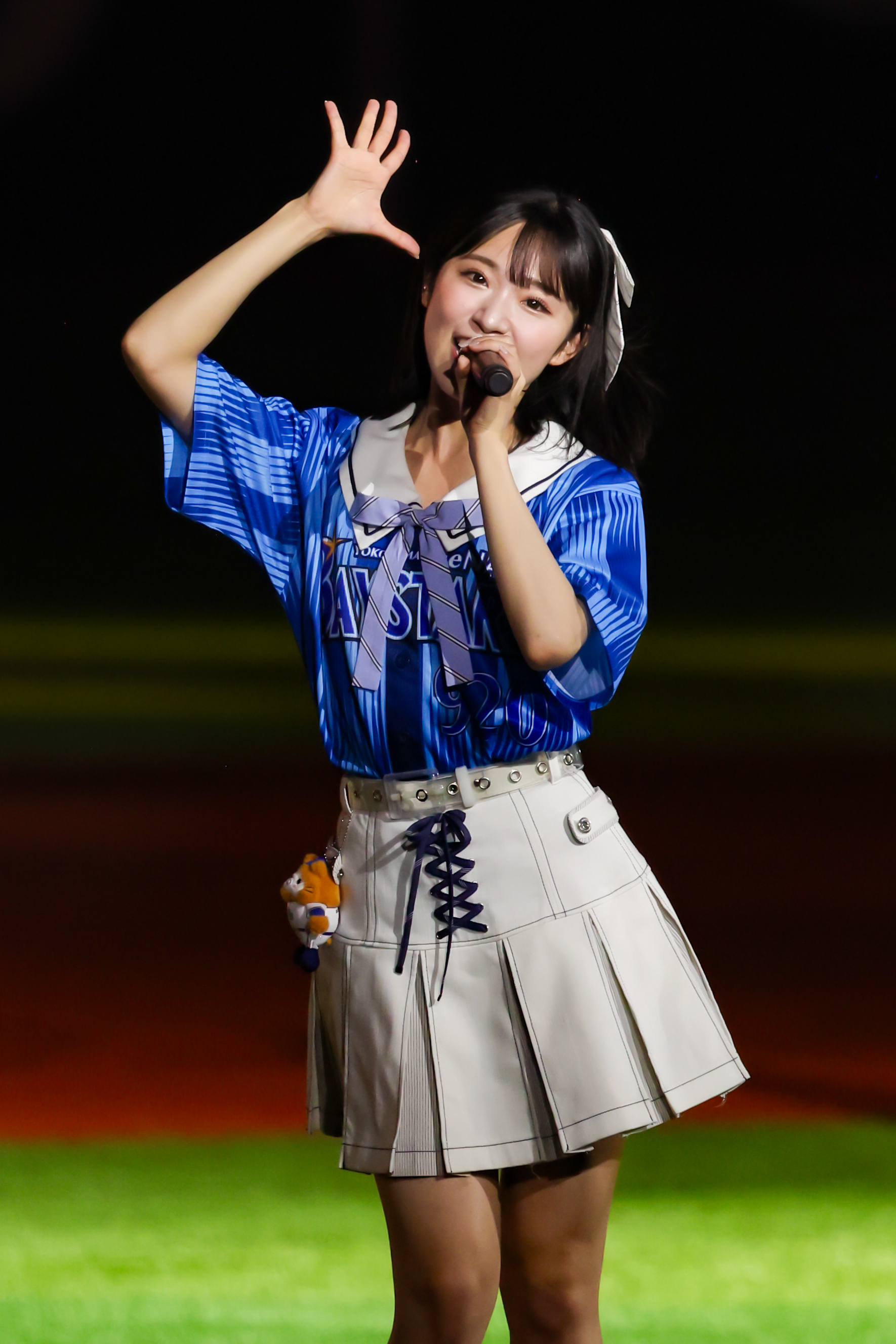 A performer wearing a blue jersey and a white skirt gestures enthusiastically while holding a microphone on stage.