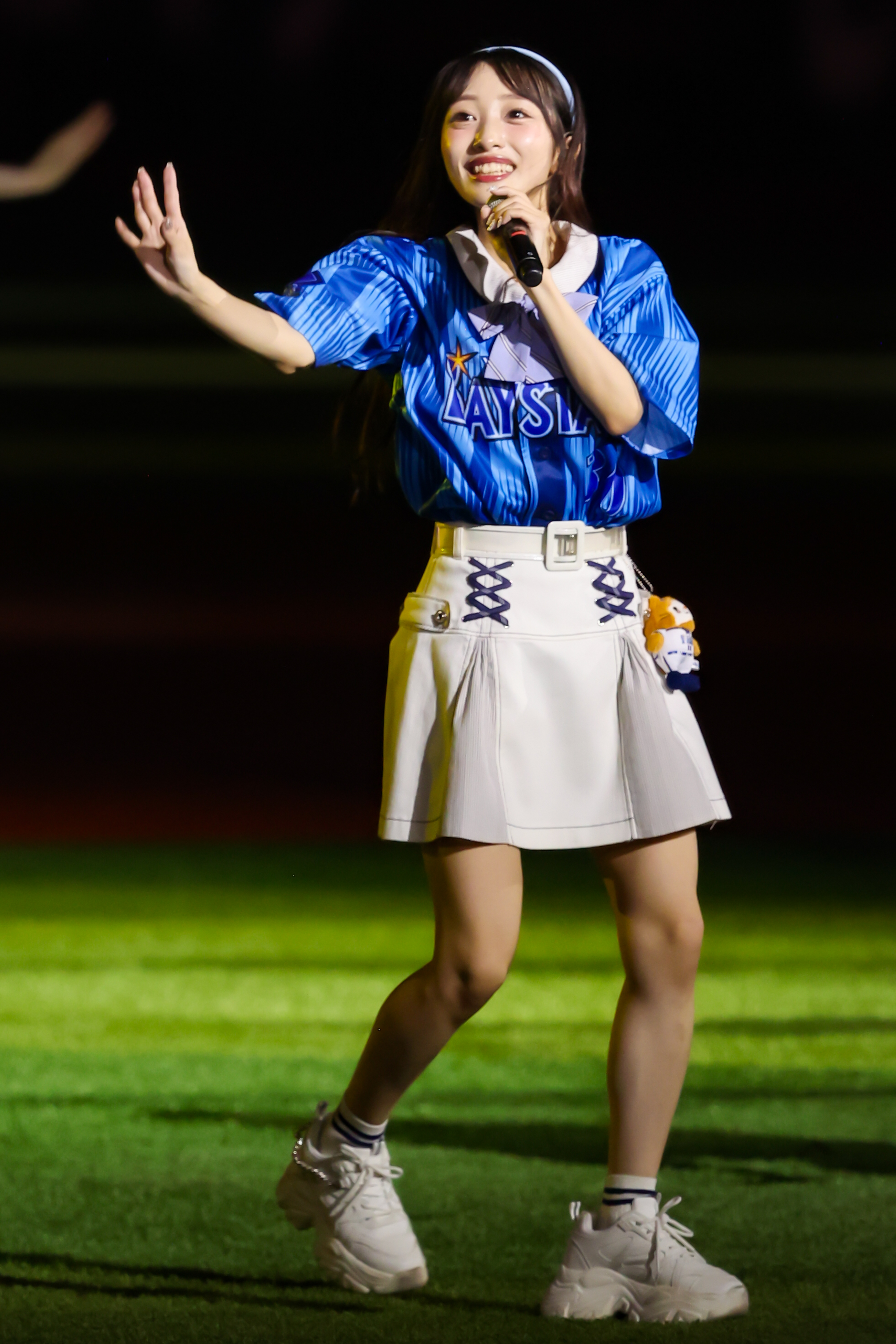 A performer in a blue jersey and white skirt is singing on stage with a microphone, smiling and gesturing with one hand.