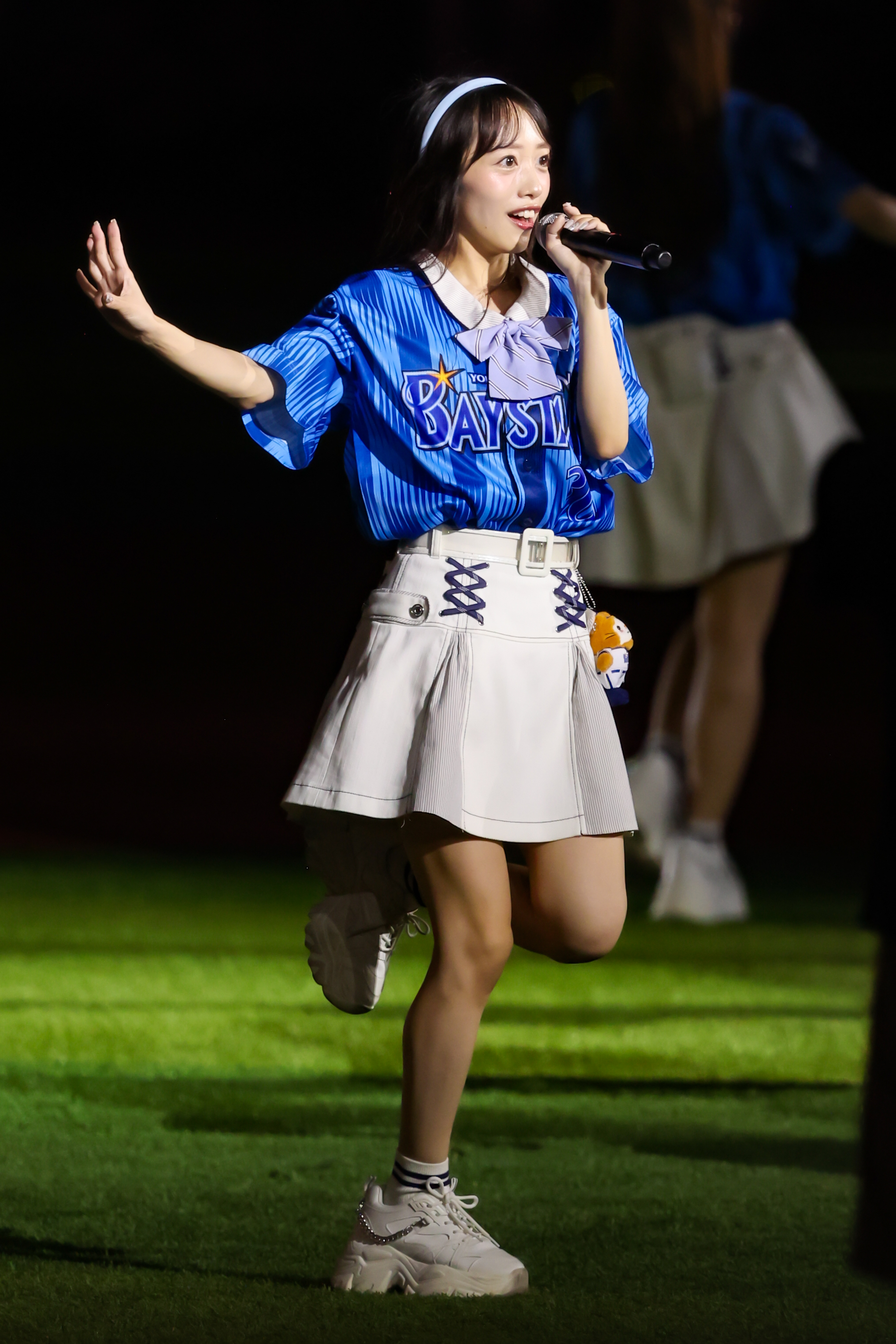 A performer in a blue jersey and white skirt energetically sings into a microphone on stage, with a blurred background of other performers.