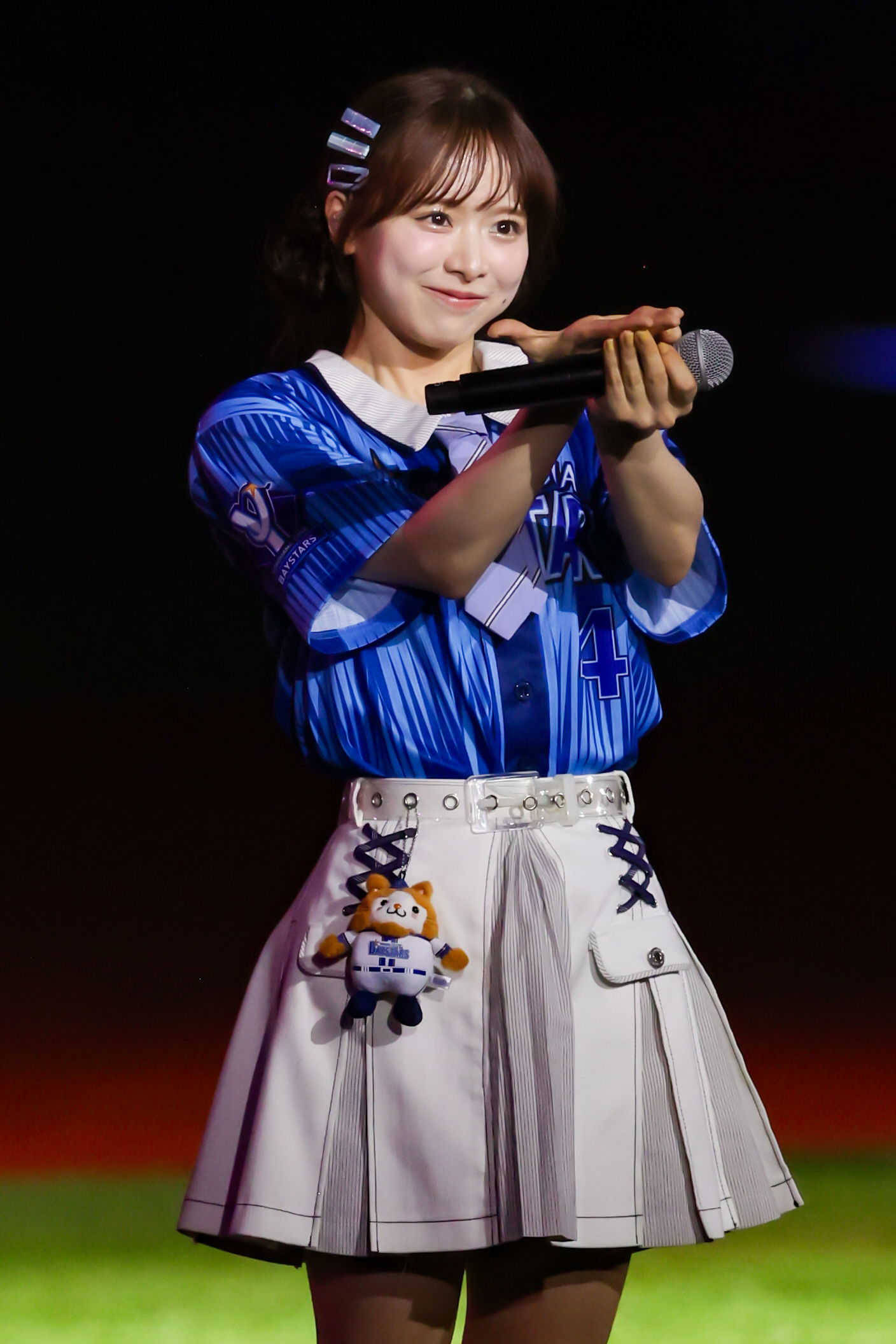 A performer in a blue sports-themed outfit holding a microphone and gesturing while smiling, with a small plush toy attached to their skirt.