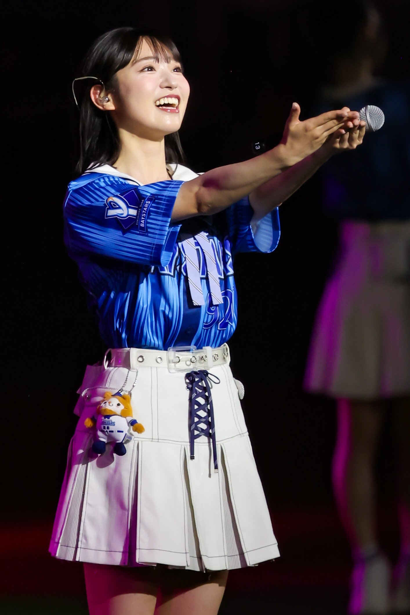 A female performer in a blue jersey and white skirt smiles and gestures while holding a microphone on stage.