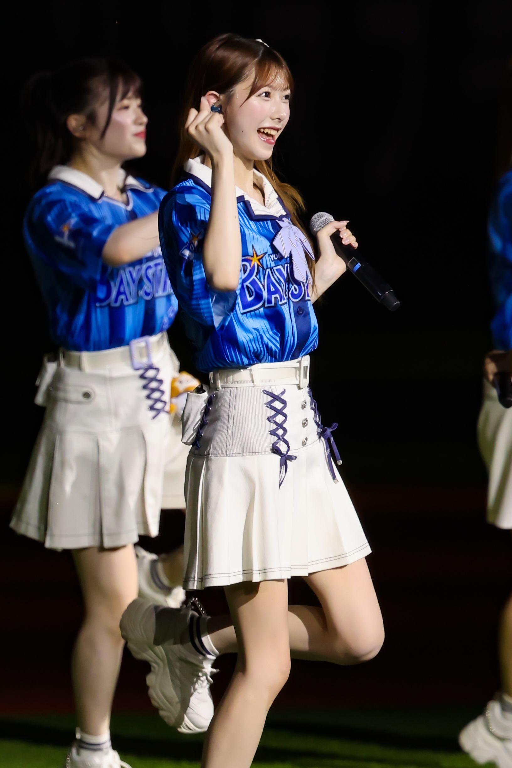 A member of AKB48 performs on stage wearing a blue shirt with 'BAYSTARS' written on it and a white skirt. She is smiling and holding a microphone, while another member is seen in the background.