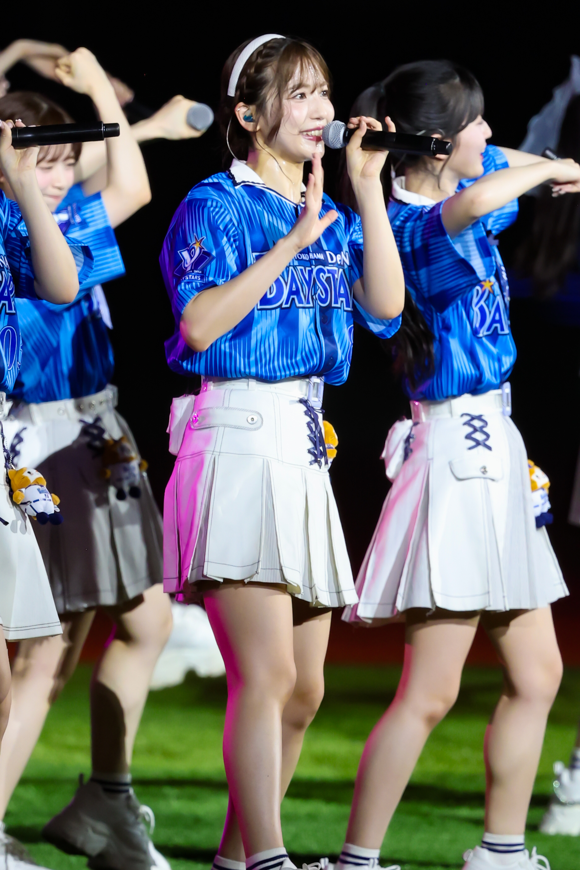 A member of AKB48 performing on stage, wearing a blue baseball jersey and a white skirt. She is holding a microphone and smiling while other members are seen behind her.