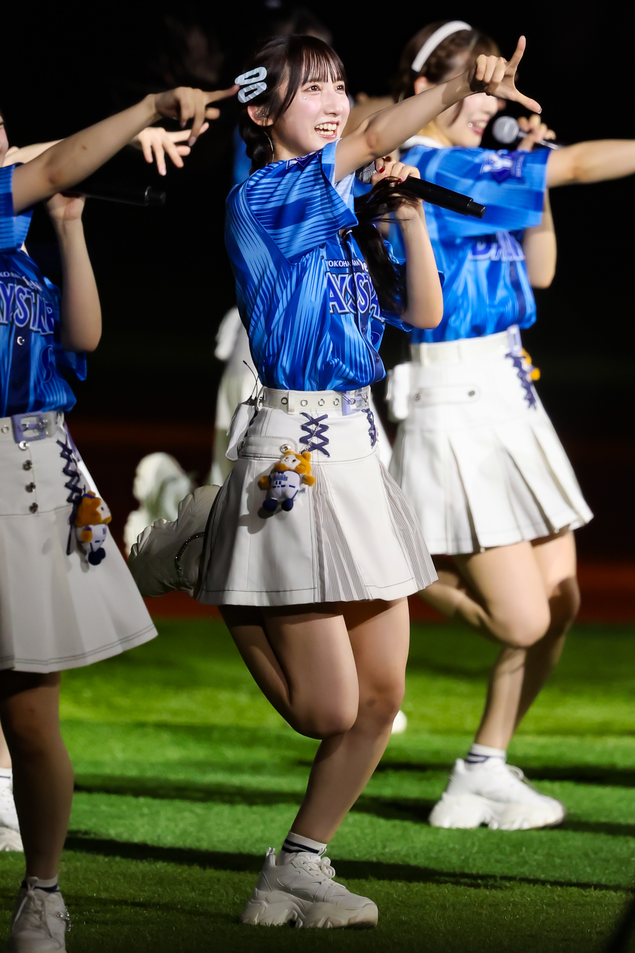 A performer from AKB48 dancing energetically on stage, wearing a blue shirt and white skirt, with a microphone in hand and a joyful expression.