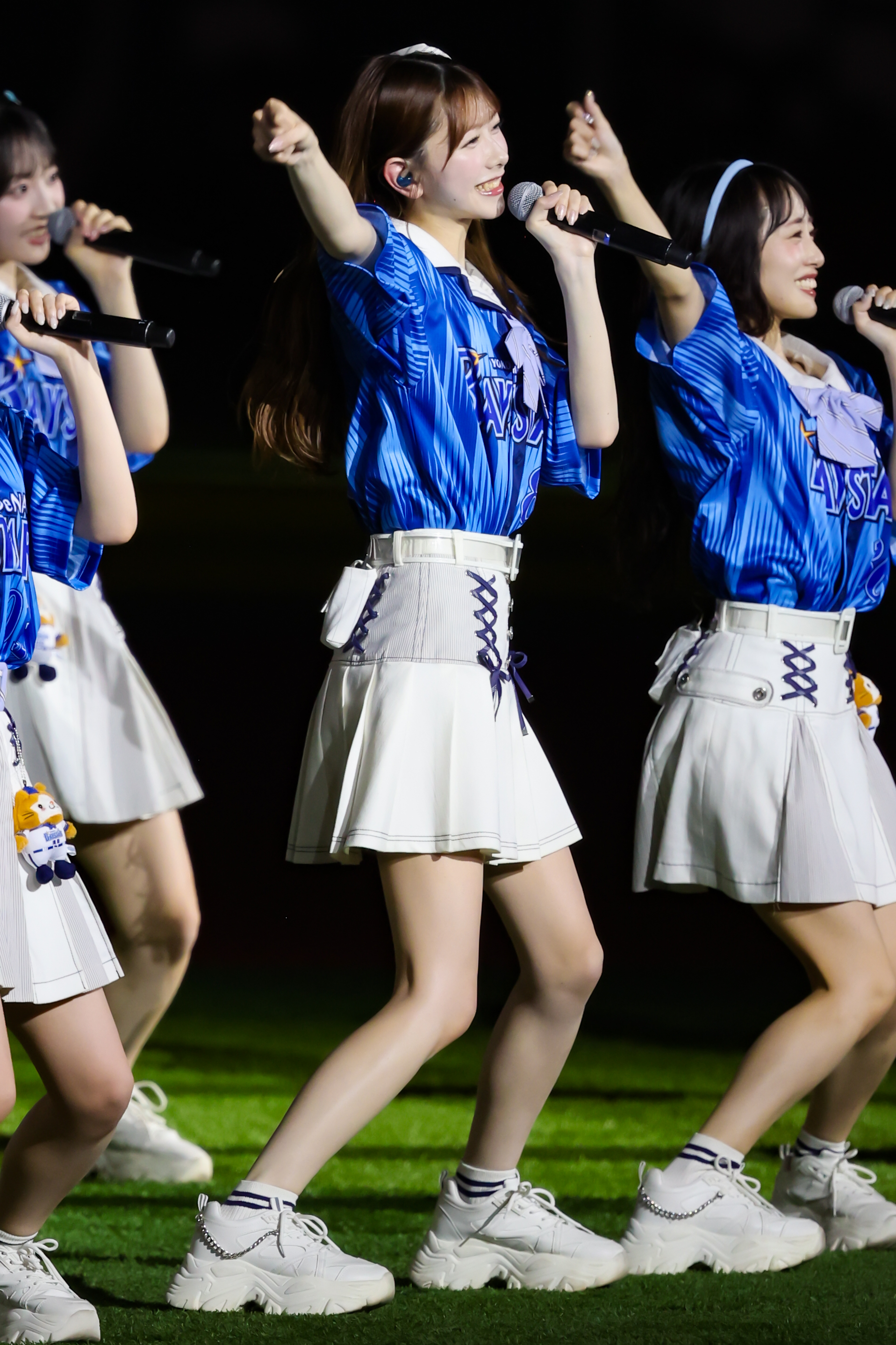 A group of performers, including a young woman in a blue shirt and white skirt, singing and dancing on stage at an outdoor event.