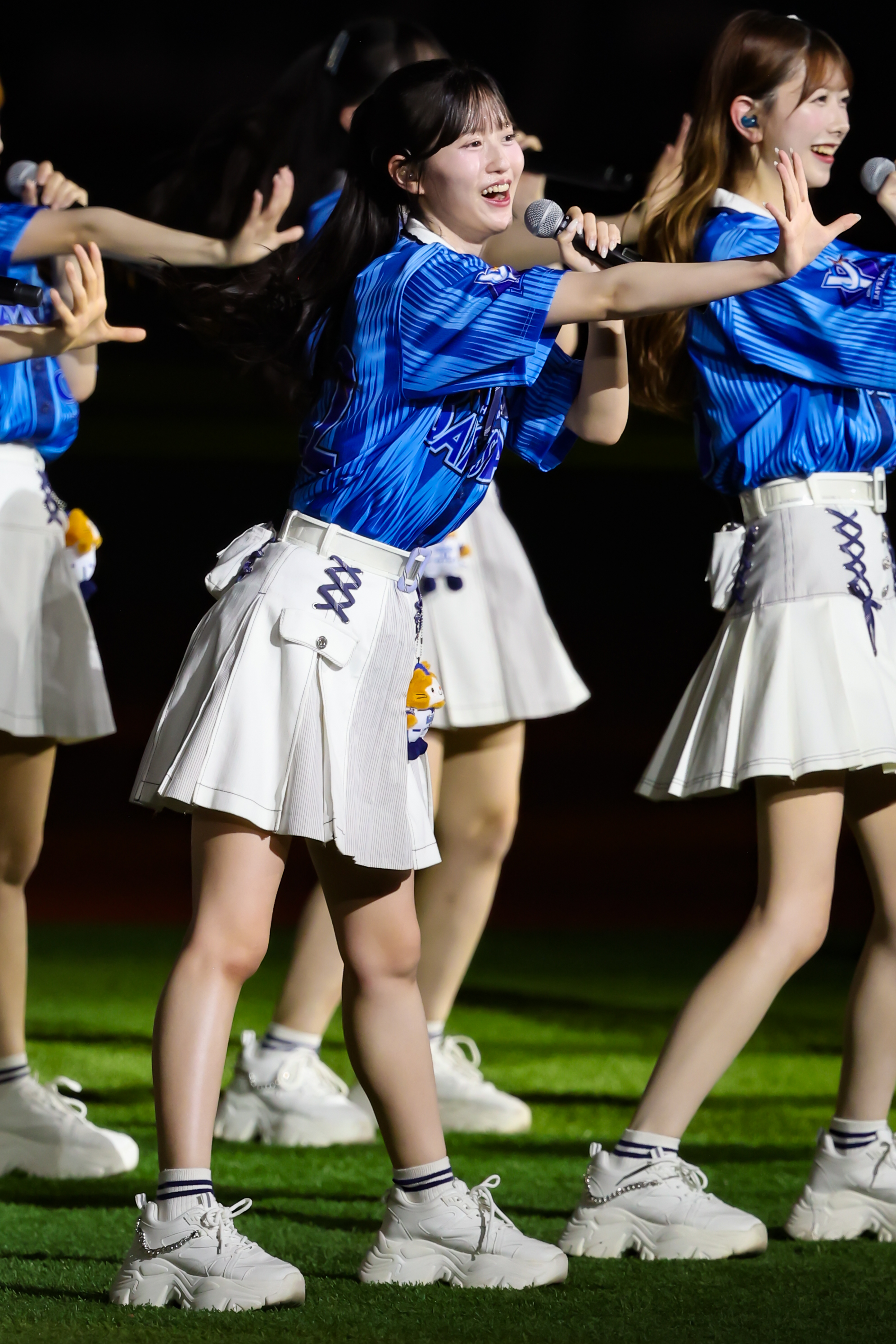 A member of AKB48 performing on stage, wearing a blue top and white skirt, holding a microphone and smiling while dancing during a concert.