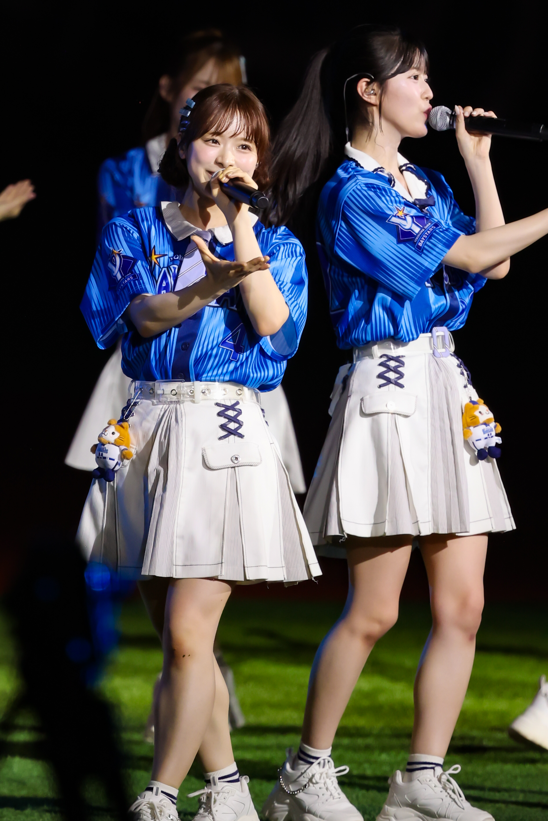 Two performers from AKB48 wearing blue shirts and white skirts, one singing into a microphone and the other holding her hand out, both smiling on stage.