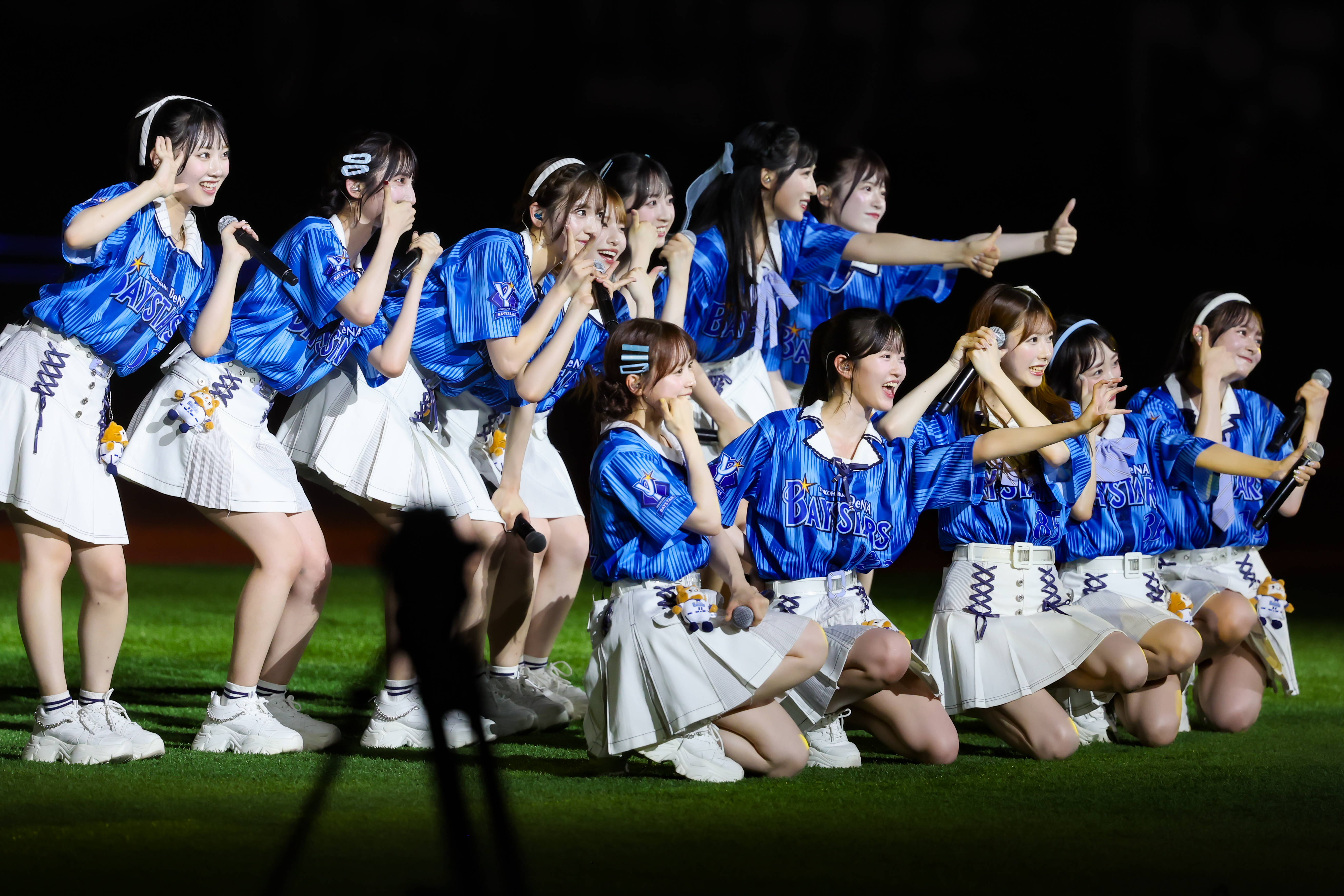 A group of performers wearing blue jerseys and white skirts, showcasing a lively performance on stage with microphones in hand.