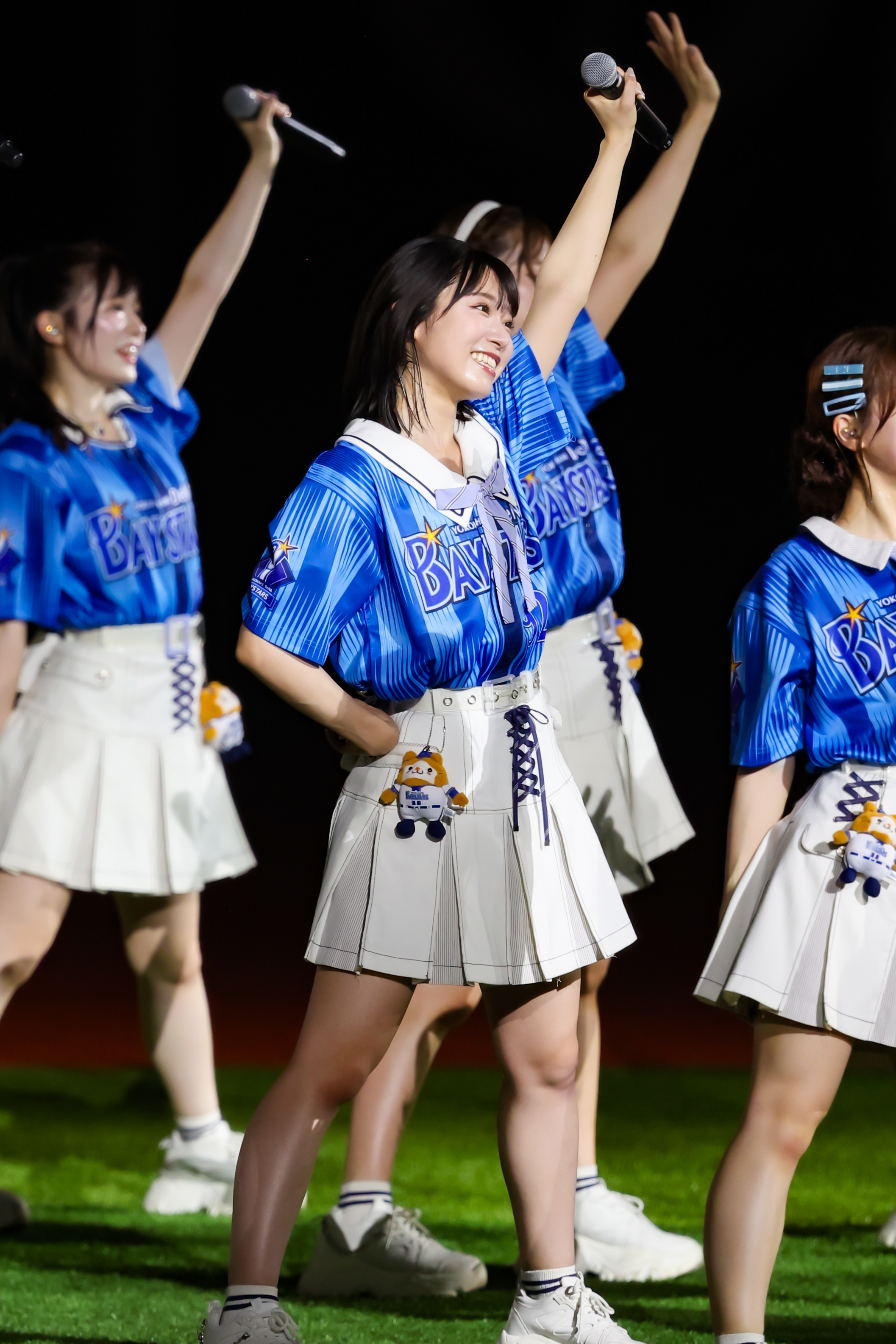 A female performer from AKB48 wearing a blue jersey and a white skirt raises a microphone while smiling on stage, with other members in the background also performing.