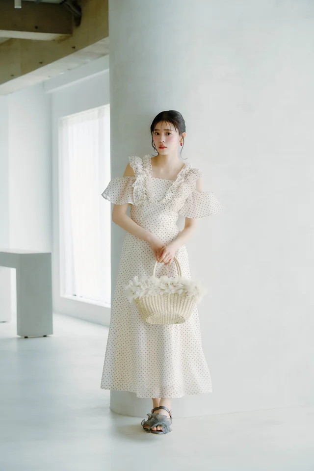女性がドット柄のフリル付きワンピースを着て、白い背景の前でかごバッグを持っている.
