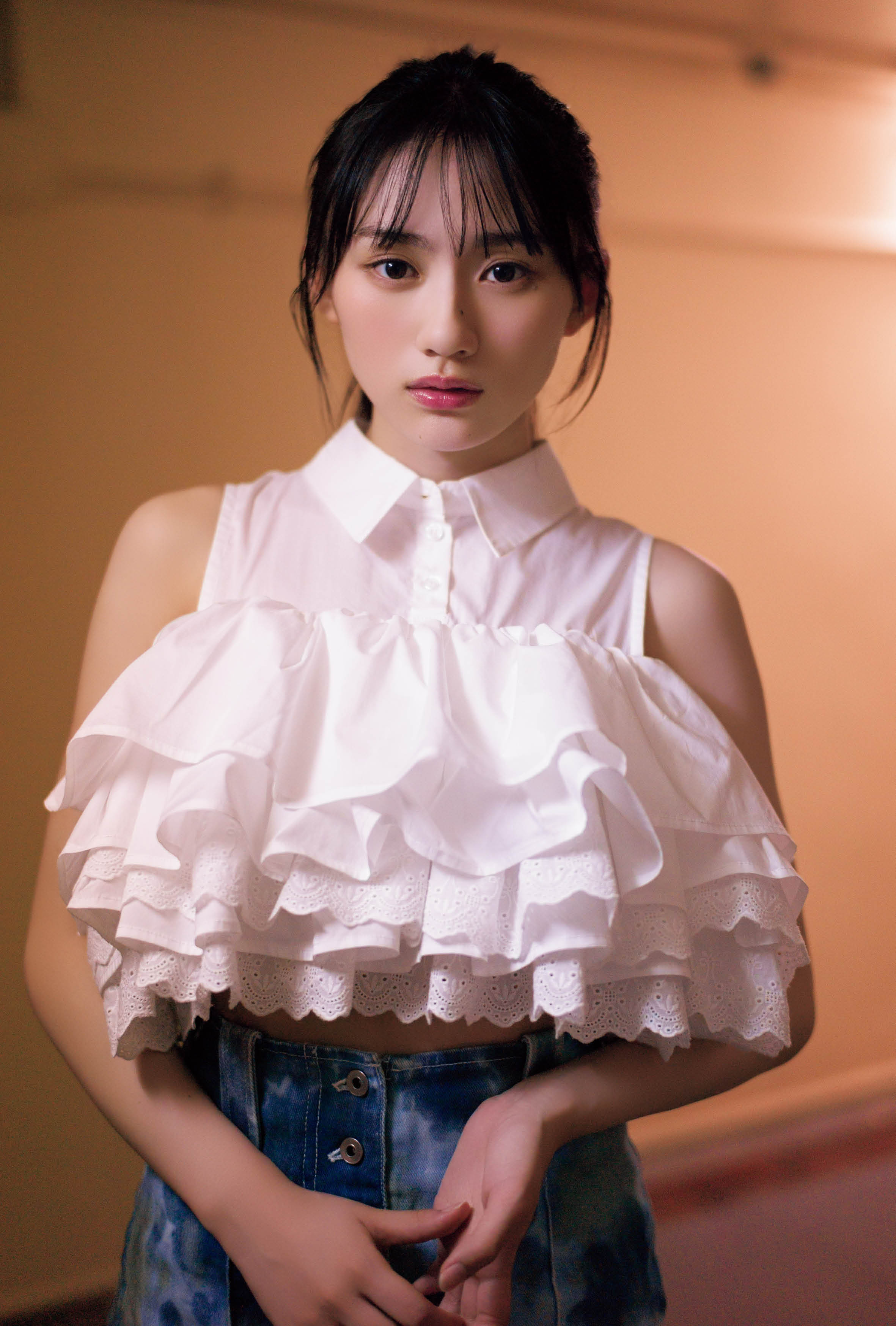 白いフリルのトップスを着た女性がカメラを見つめている写真。彼女は青いジーンズを合わせており、柔らかな照明の中で撮影されている。