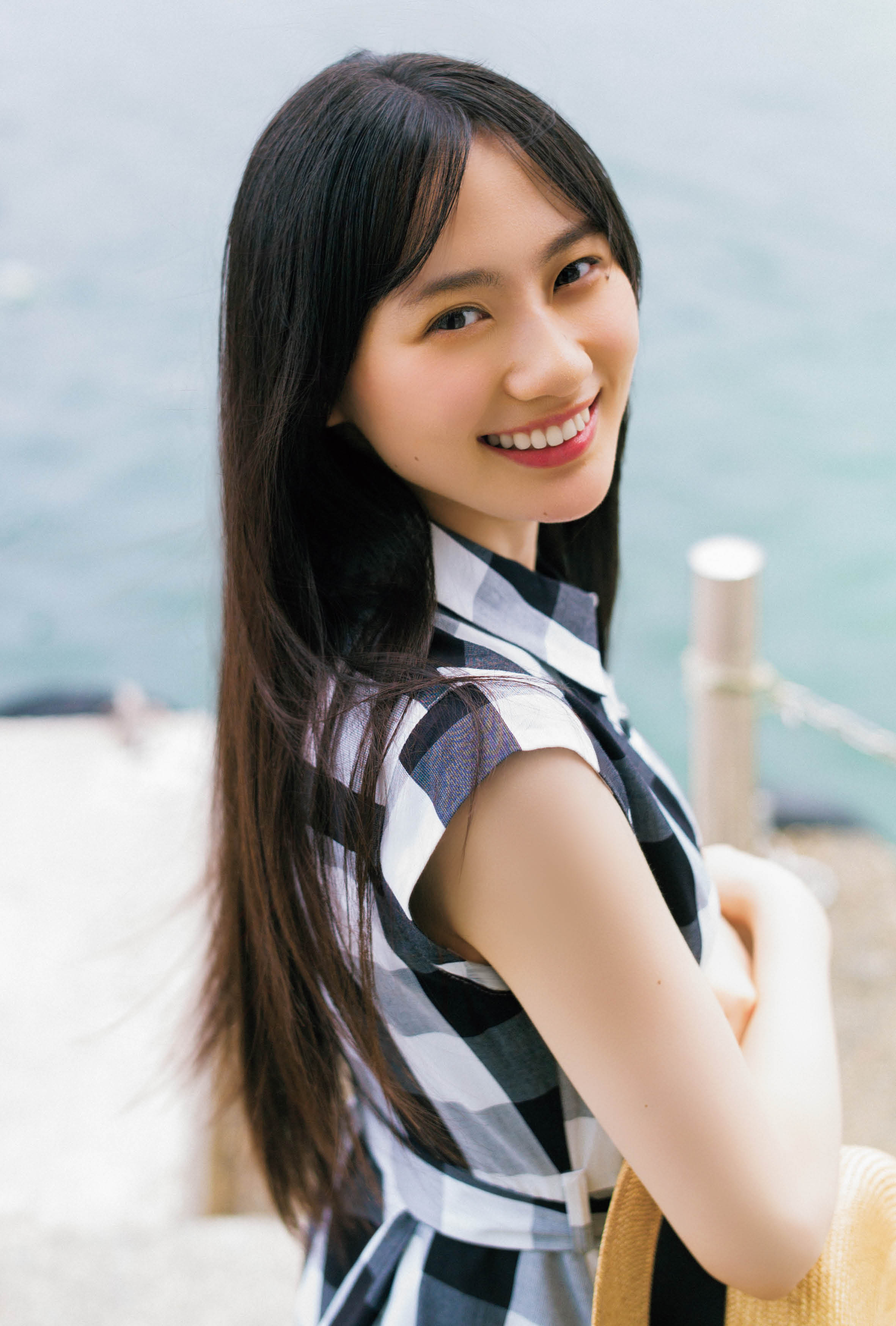 海辺で微笑む女性、チェック柄の服を着てカメラに向かっている。背景には青い海が広がっている。