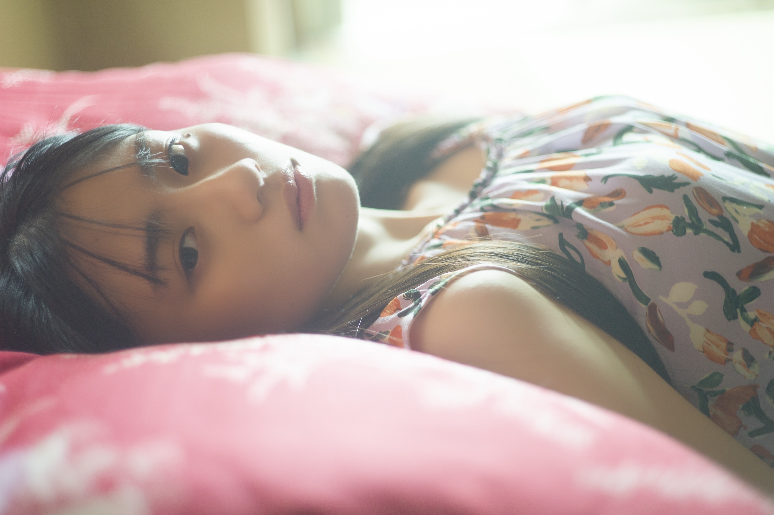 穏やかな表情の女性が布団の上に横たわっている。彼女は花柄の衣服を着ており、静かな眼差しを向けている。柔らかな光に包まれた穏やかな雰囲気を醸し出している。