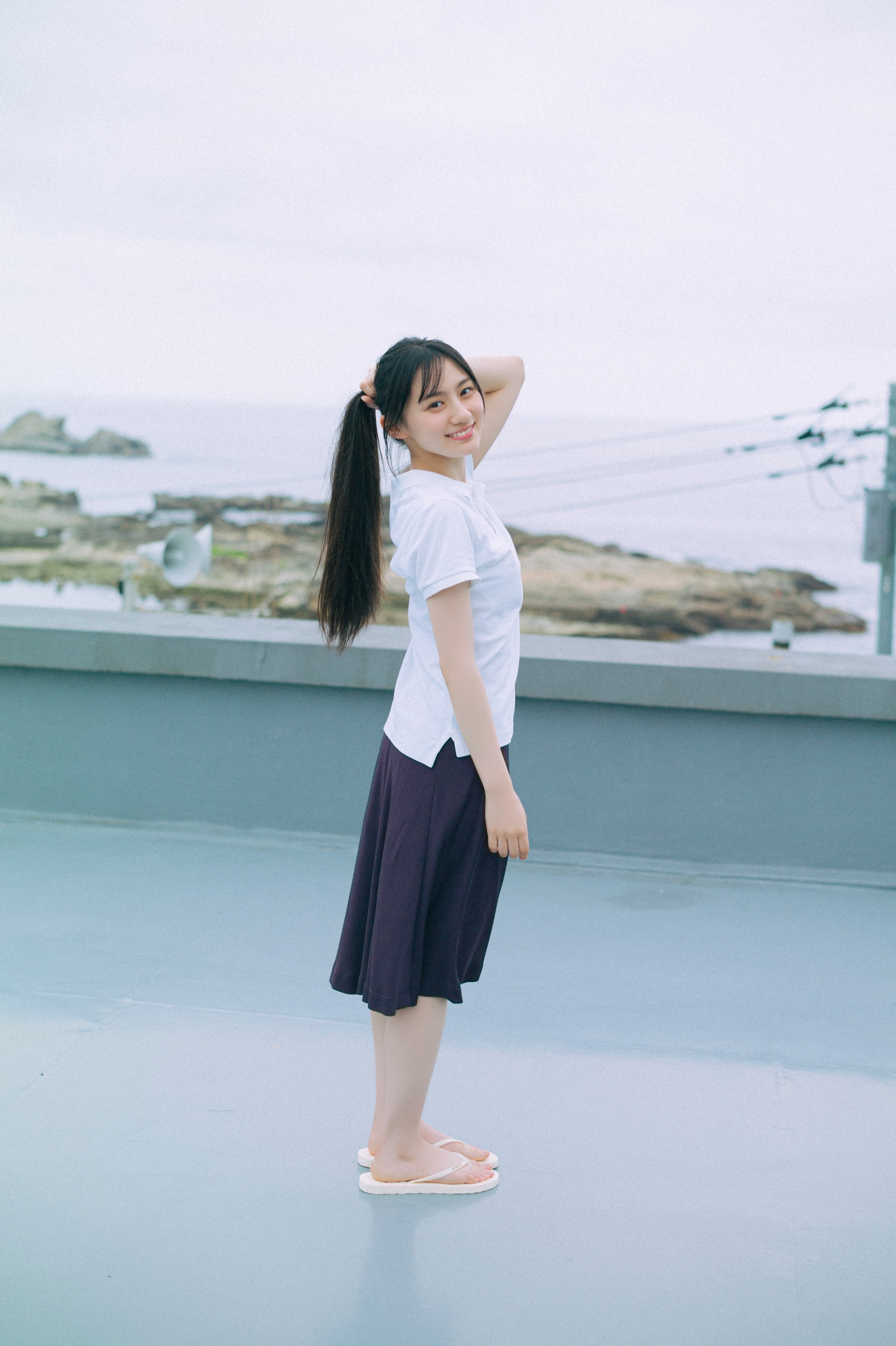 海辺で笑顔を見せる女性が、白いシャツと黒いスカートを着て立っている画像。