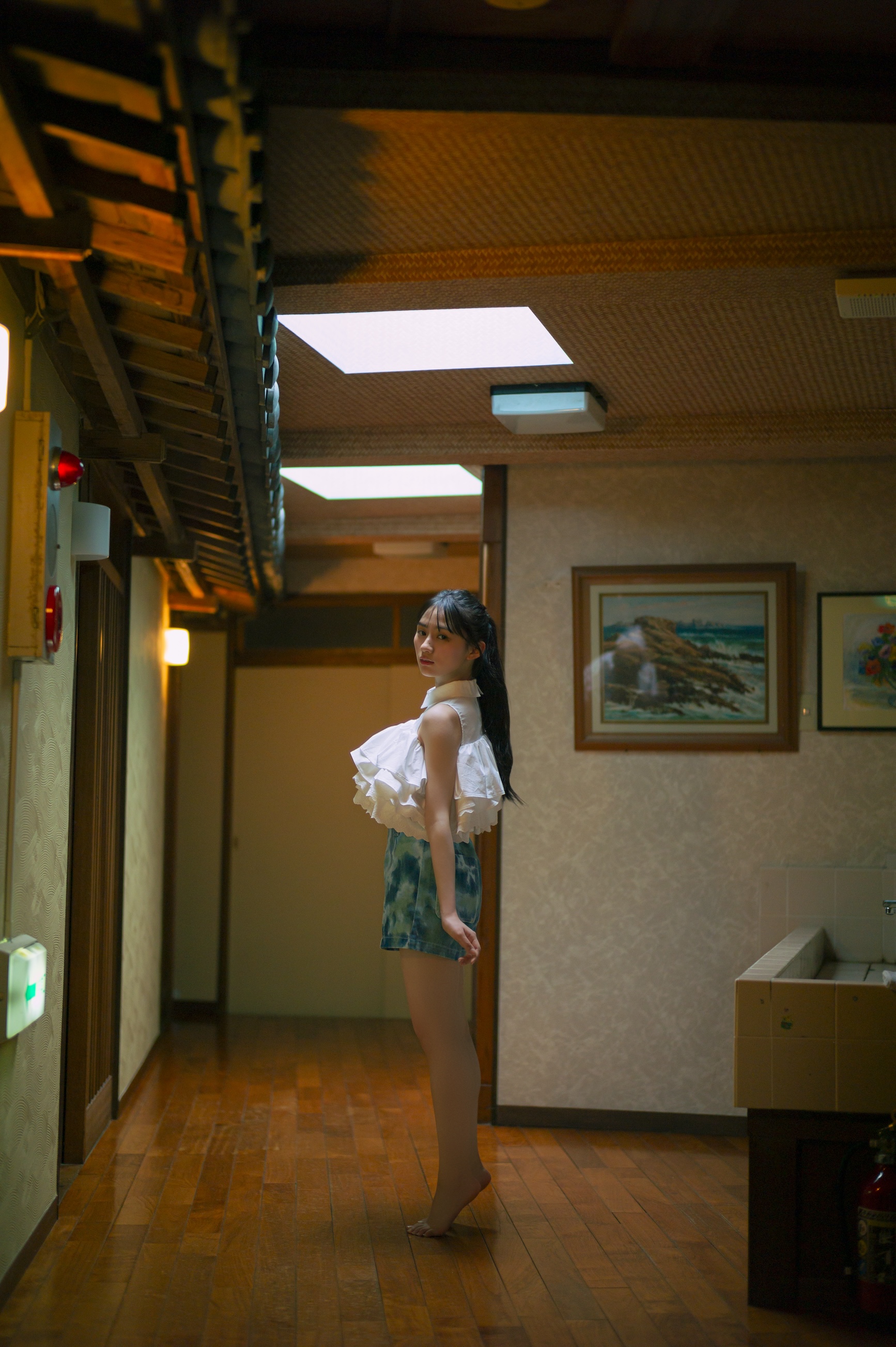 海辺の旅館の廊下でポーズを決める女性。白いフリルのトップスと青いショートパンツを着用し、静かな表情を見せている。