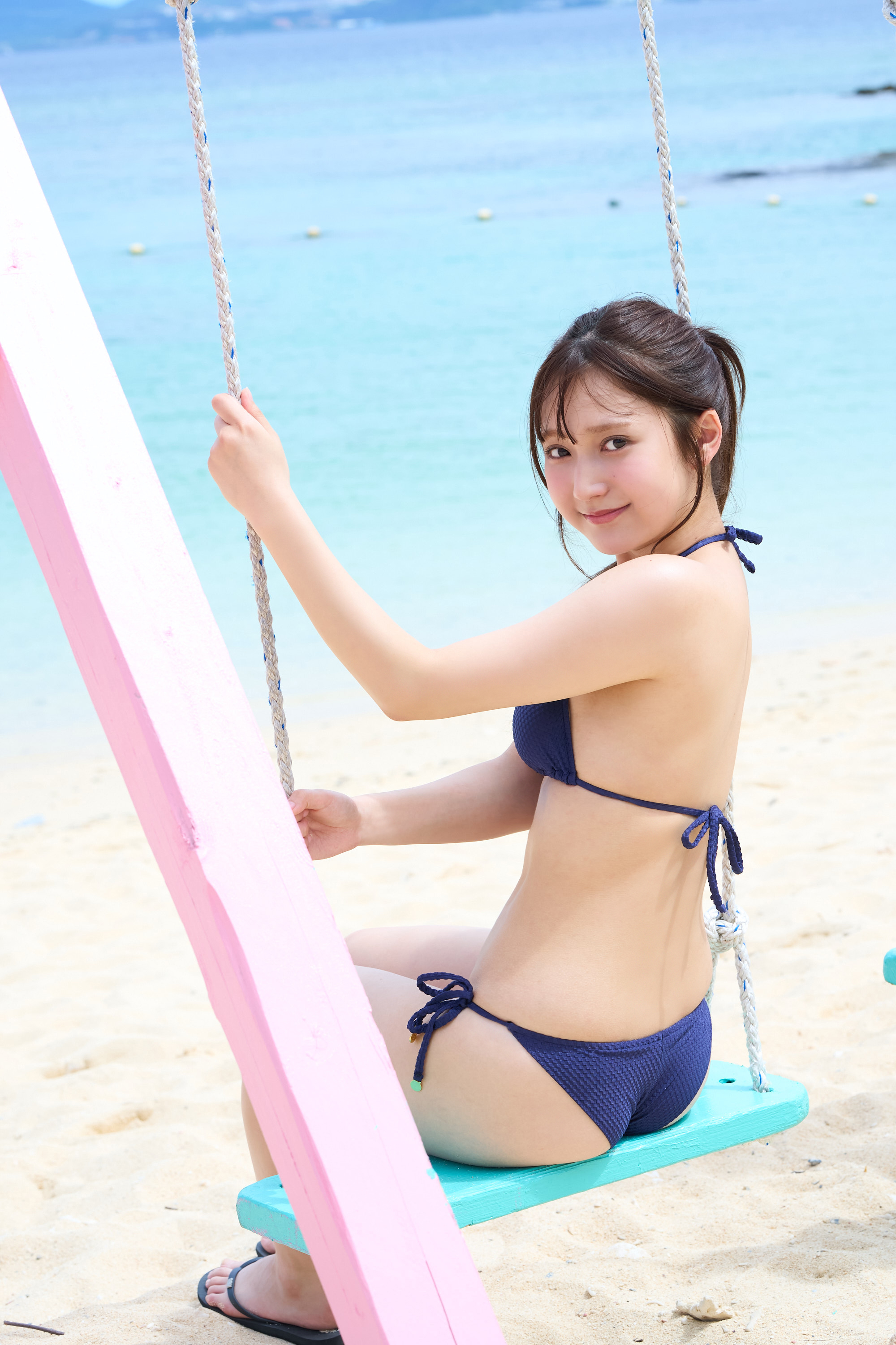 海辺のブランコに座る女性が、青い水着を着て微笑んでいる写真