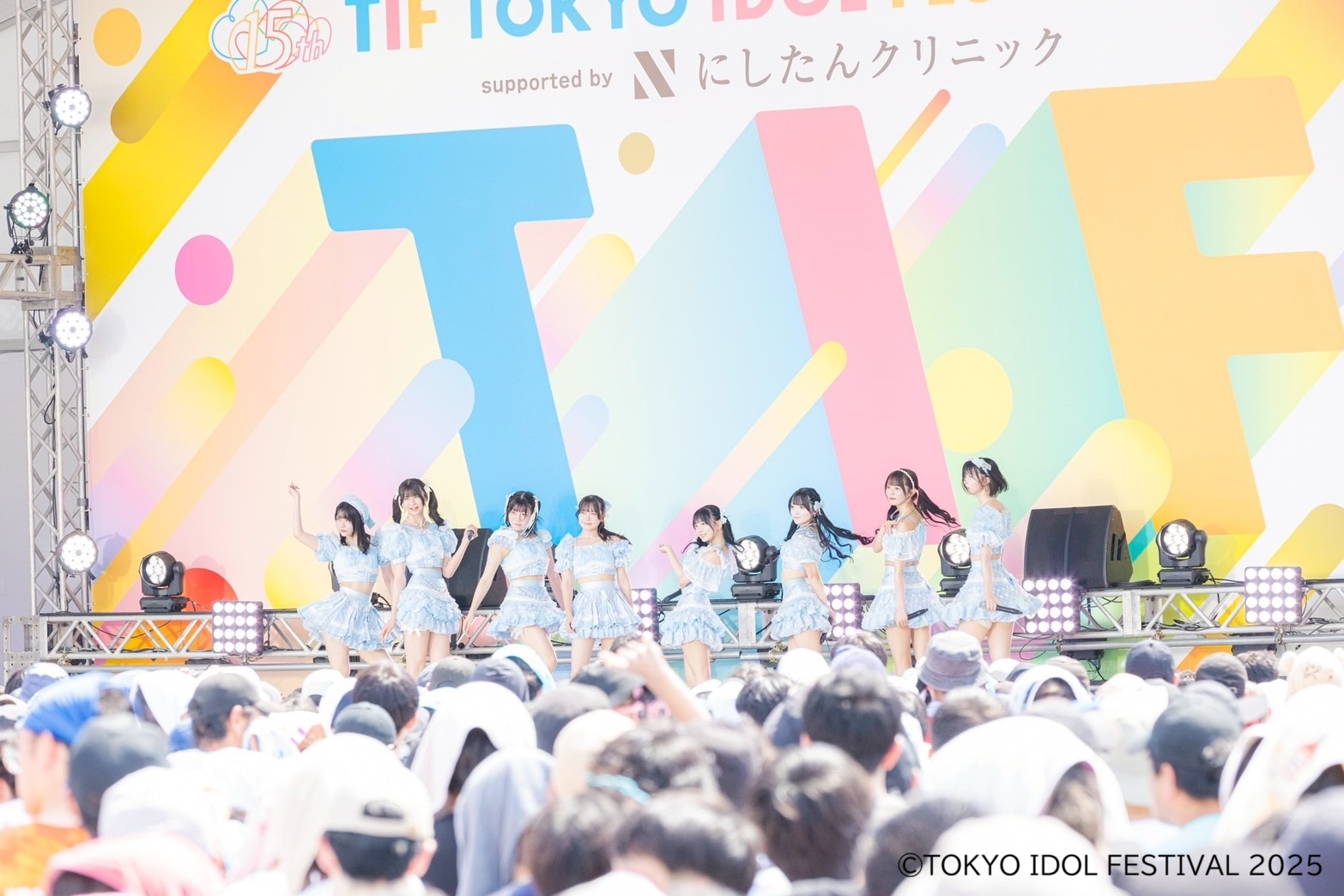 アイドルグループがパフォーマンスを行っている様子。カラフルな背景の前で、衣装を着たメンバーがステージで踊り、観客が見守っている。